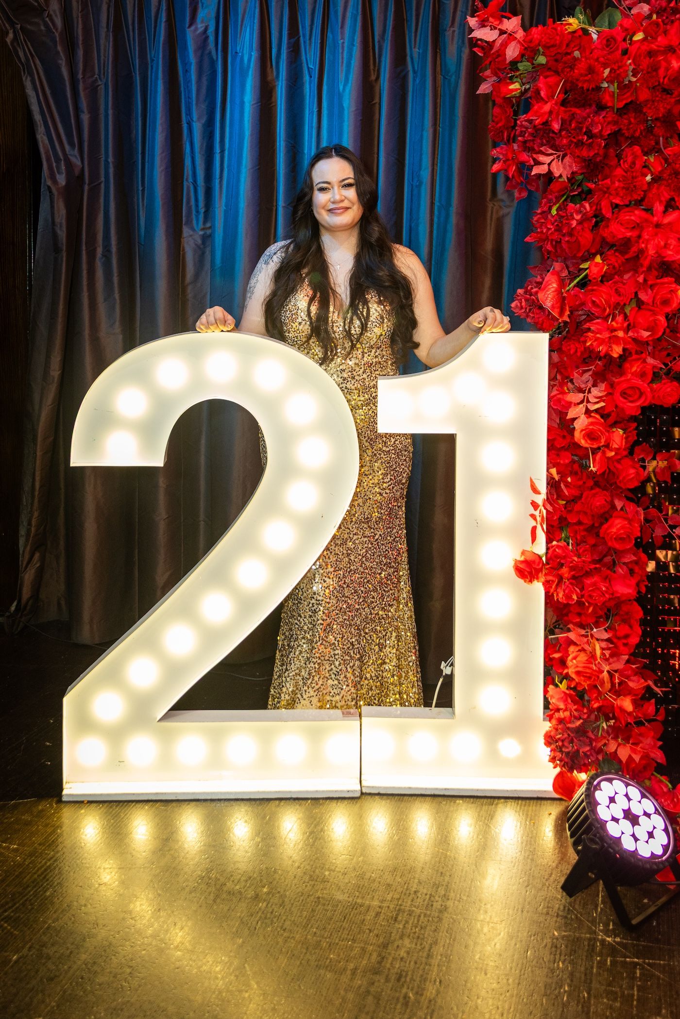Woman in gold sequin gown posing with illuminated marquee number 21 sign at birthday celebration with red floral backdrop.