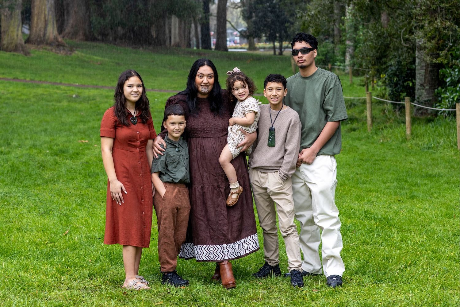 Family of six posing together on green grass in a park, smiling for a outdoor portrait photo.