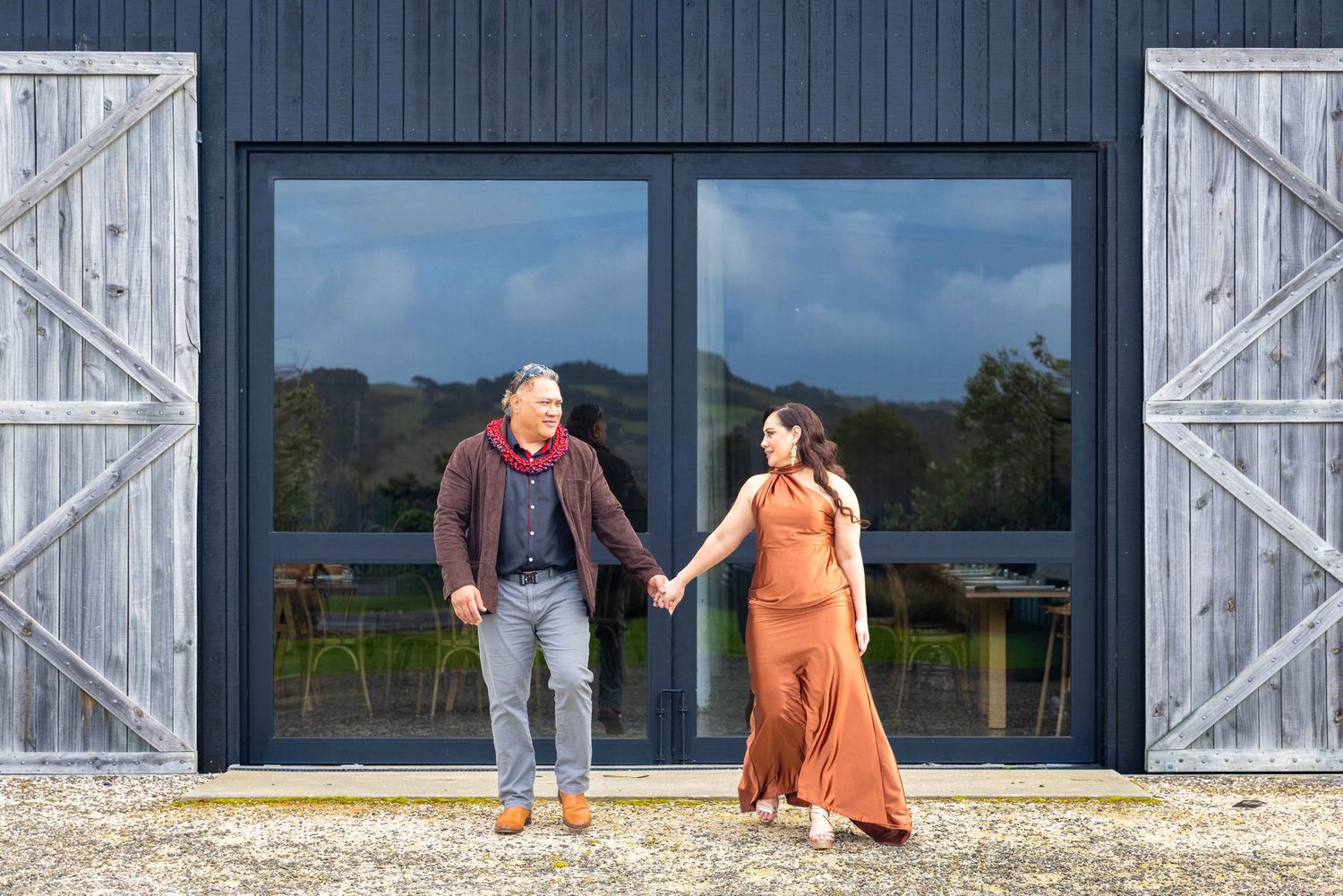 Couple holding hands walking in front of a dark barn building with wooden doors outdoors.