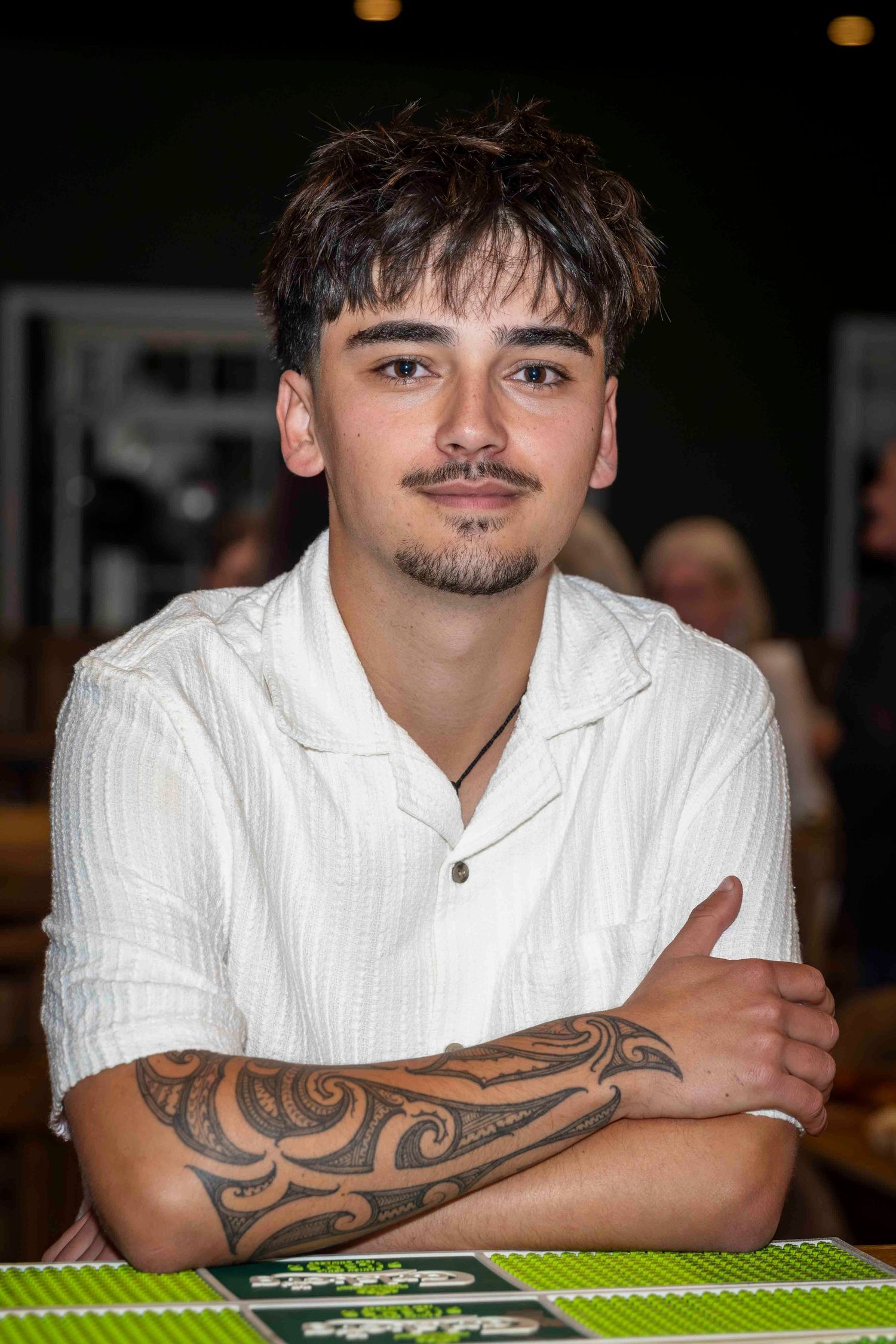 Young man with tribal tattoo on arm sits cross-armed, wearing white shirt, smiling at camera indoors.