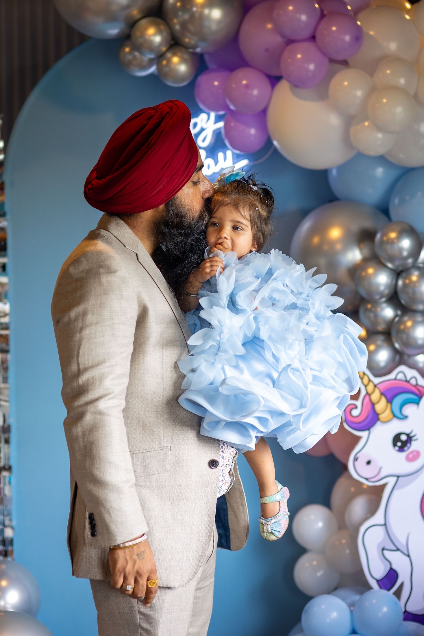 A Sikh father in a red turban and beige suit kisses his baby girl in a blue ruffled dress at a unicorn birthday party.