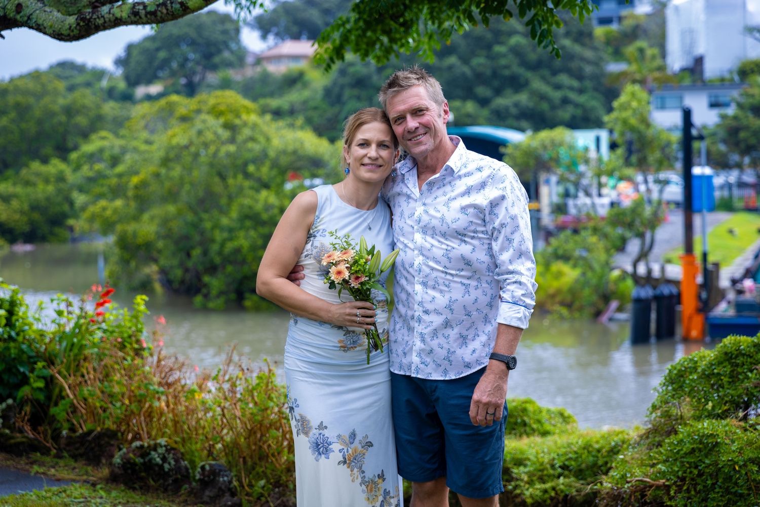 Smiling couple posing outdoors near a river, woman holding a floral bouquet in a white dress, man in a floral shirt.