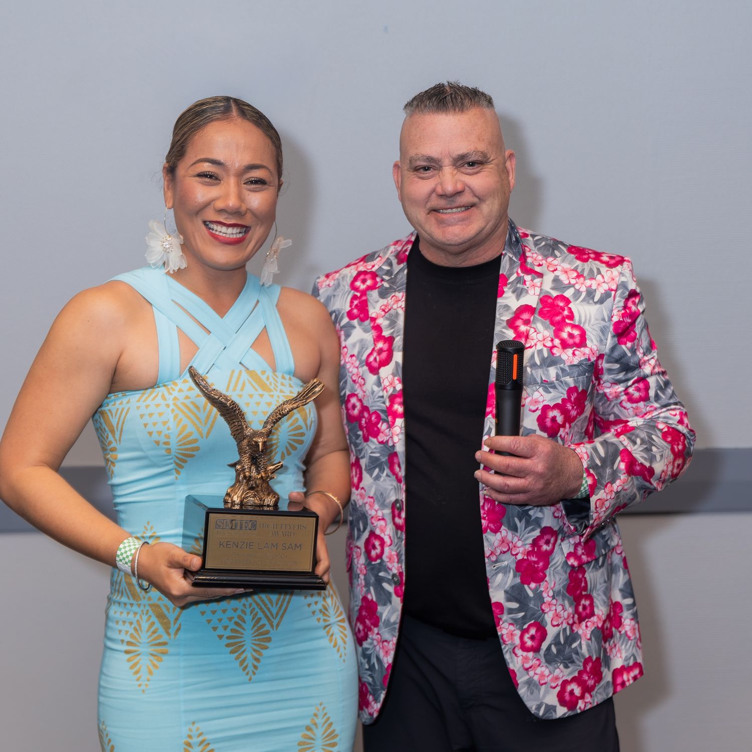 Woman in blue dress holds eagle trophy beside man in floral blazer at awards ceremony.