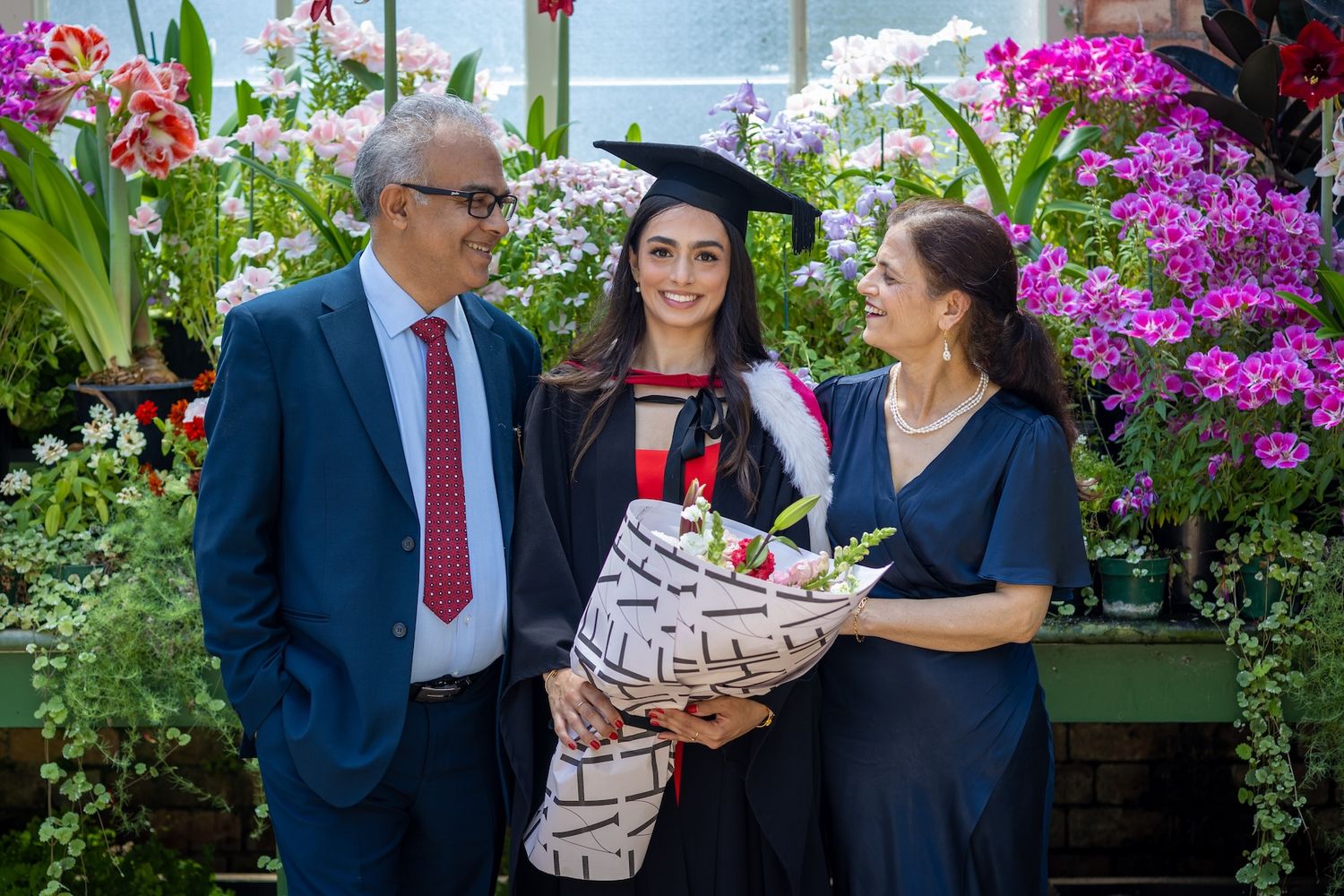 Graduate in cap and gown poses with proud parents among colorful flowers and orchids.