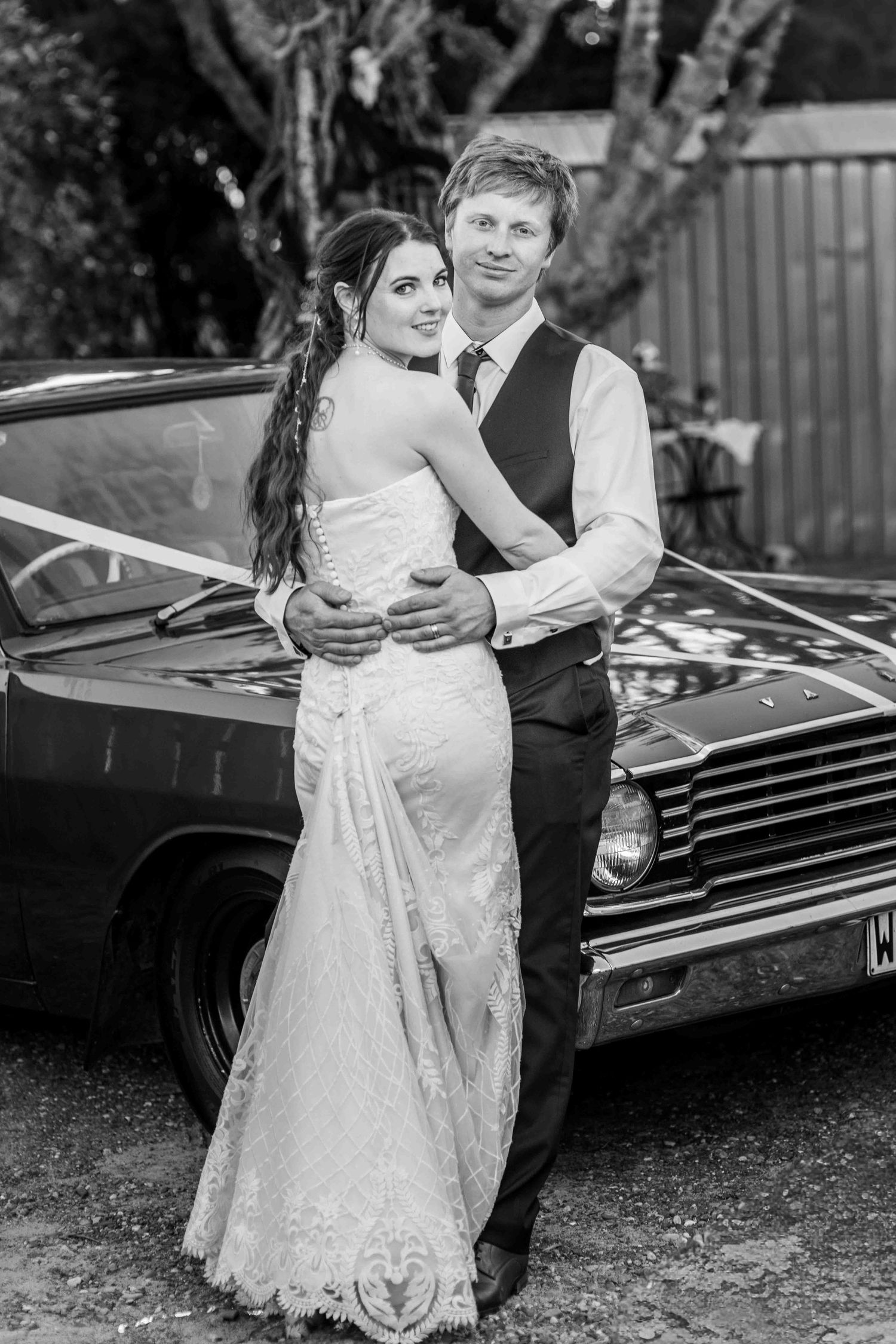 Black and white photo of a bride and groom embracing in front of a classic car on their wedding day.
