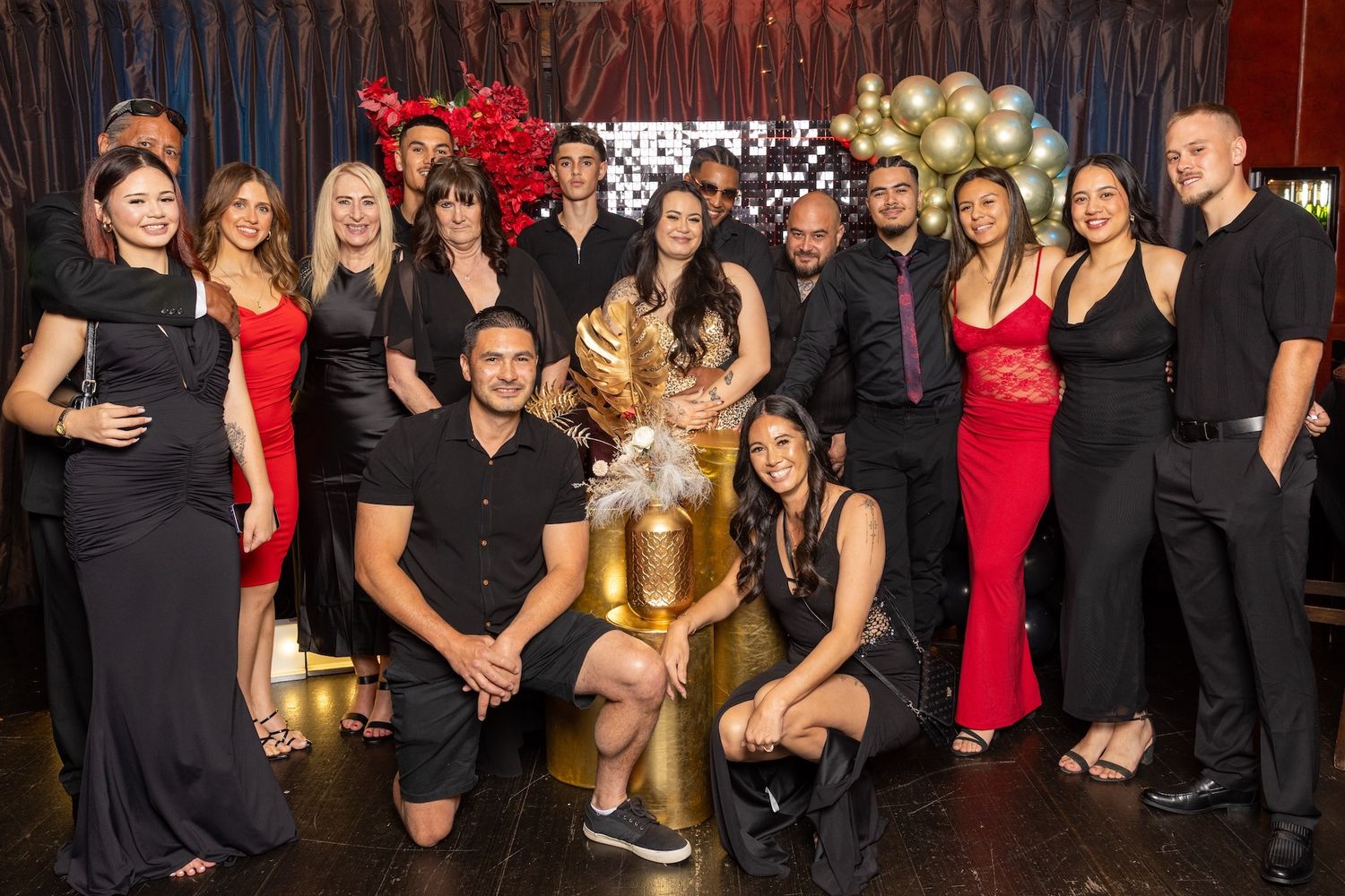 Group of elegantly dressed people posing together at a festive party with gold and red decorations.