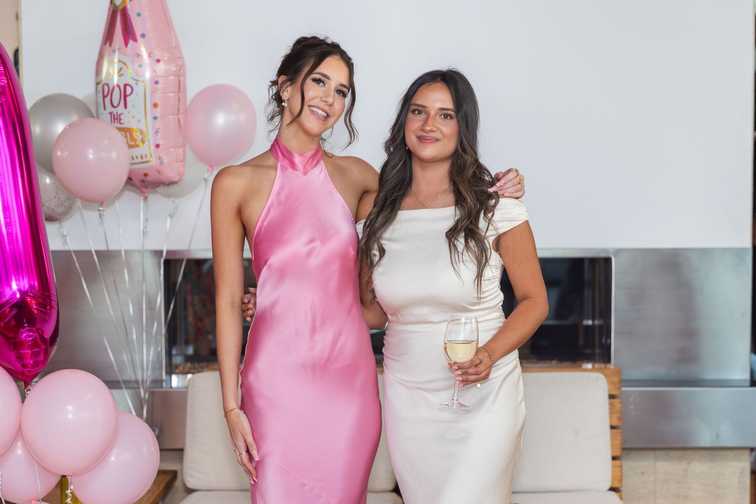Two women pose together at a pink-themed baby shower, one in a pink satin dress and one in a white dress.