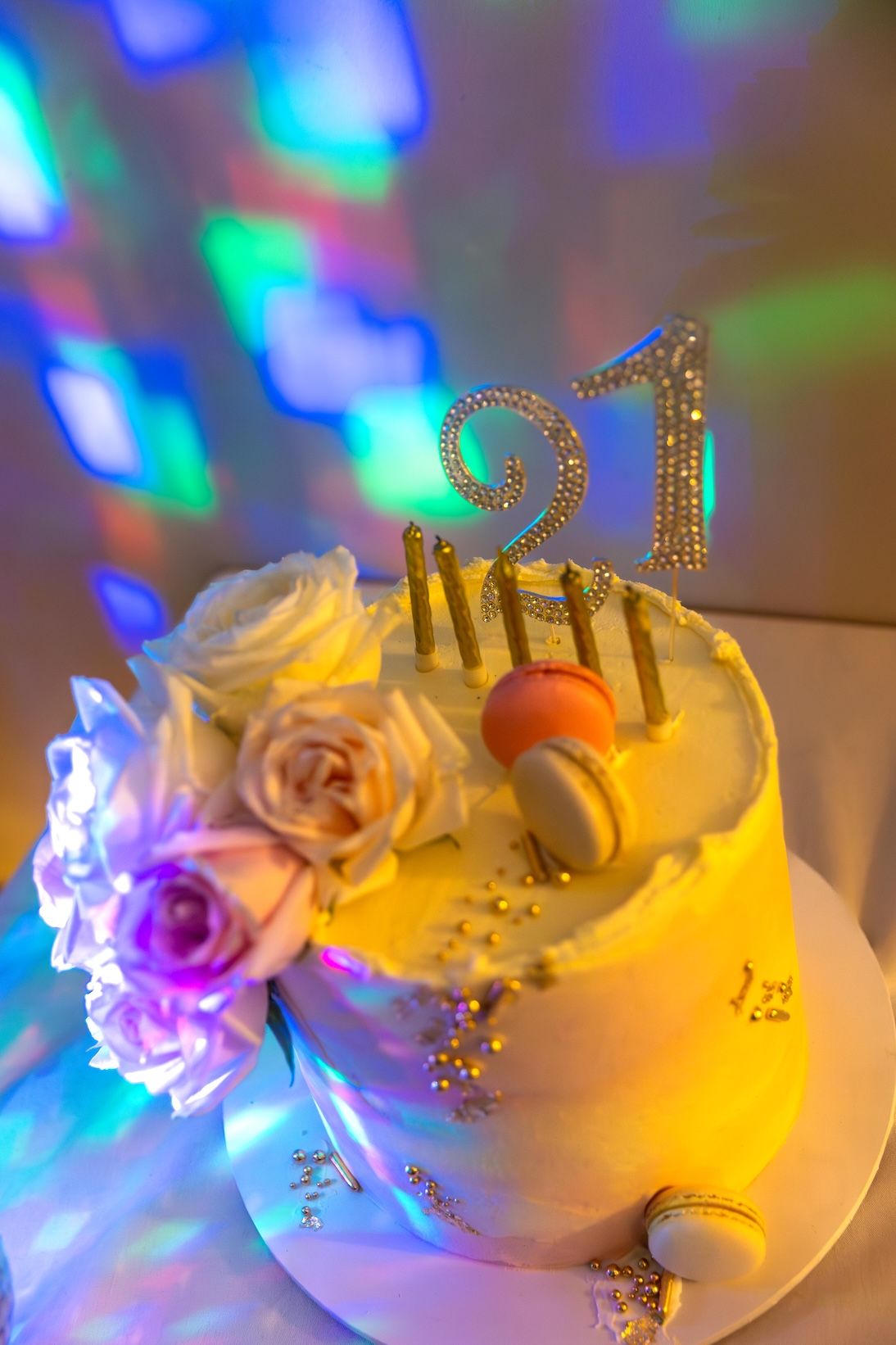 A glittery 21st birthday cake with white frosting, roses, macarons, and colorful bokeh heart lights in the background.
