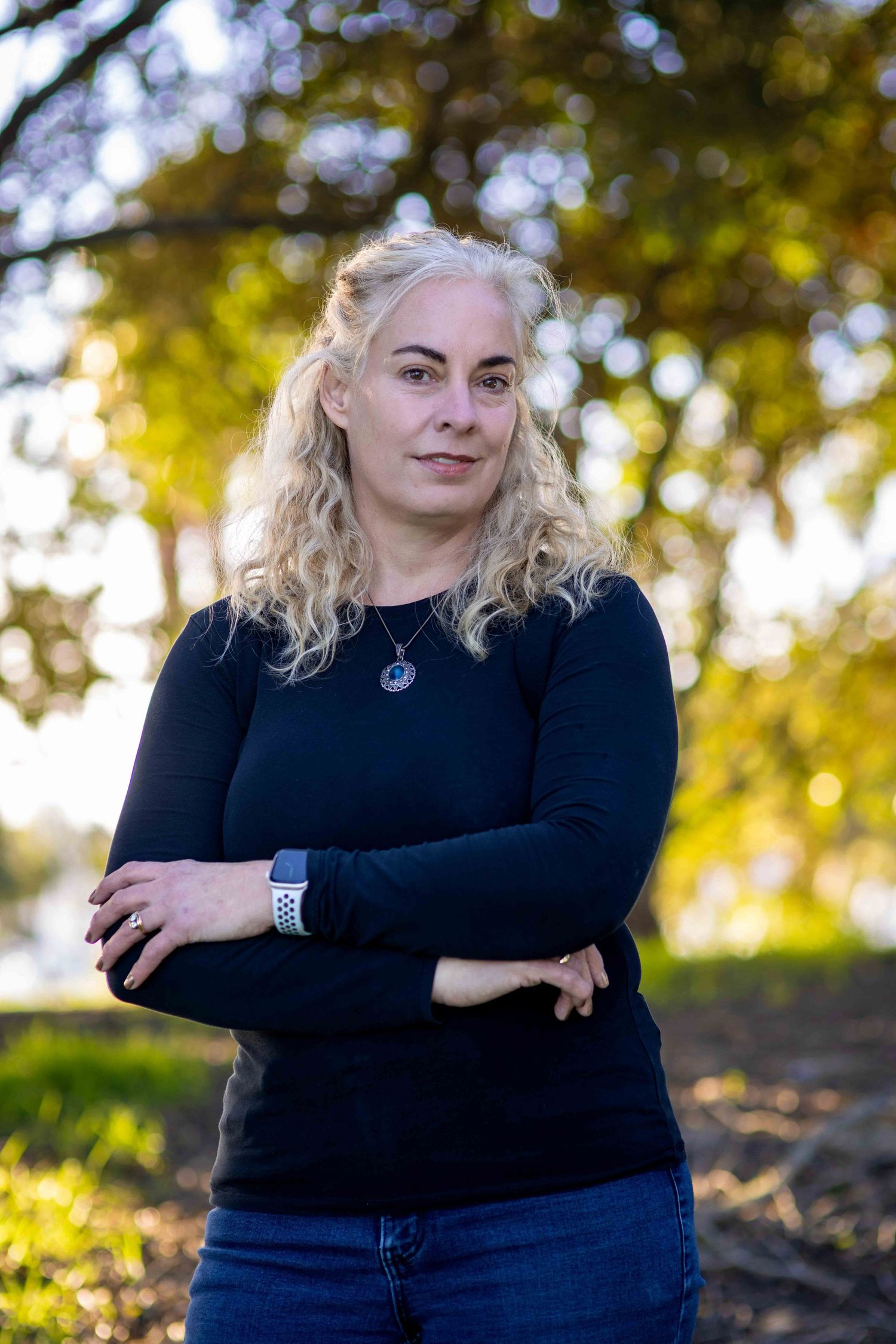 Woman with blonde curly hair in black long-sleeve top stands outdoors with arms crossed, bokeh trees background.