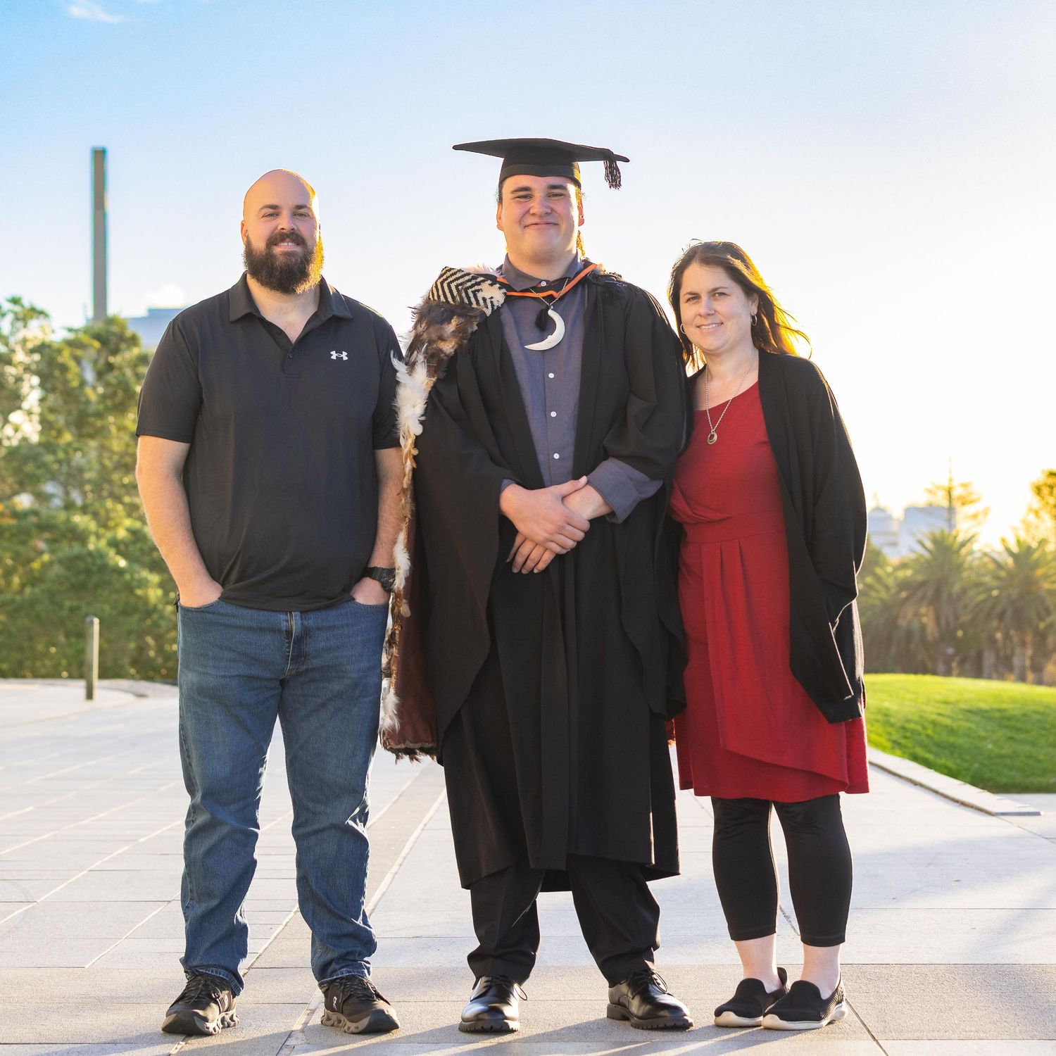 Graduate in cap and gown poses with two proud family members outdoors at a university graduation ceremony.
