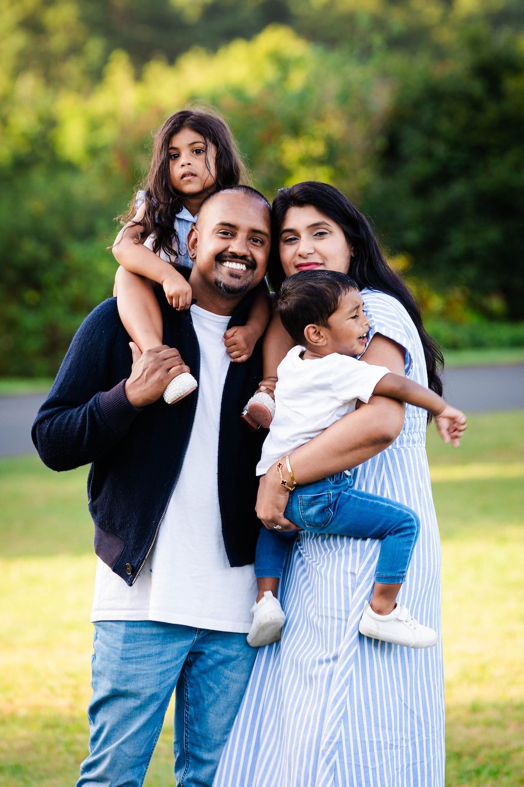 Happy South Asian family of four posing outdoors, parents holding two young children, smiling in a lush green park.