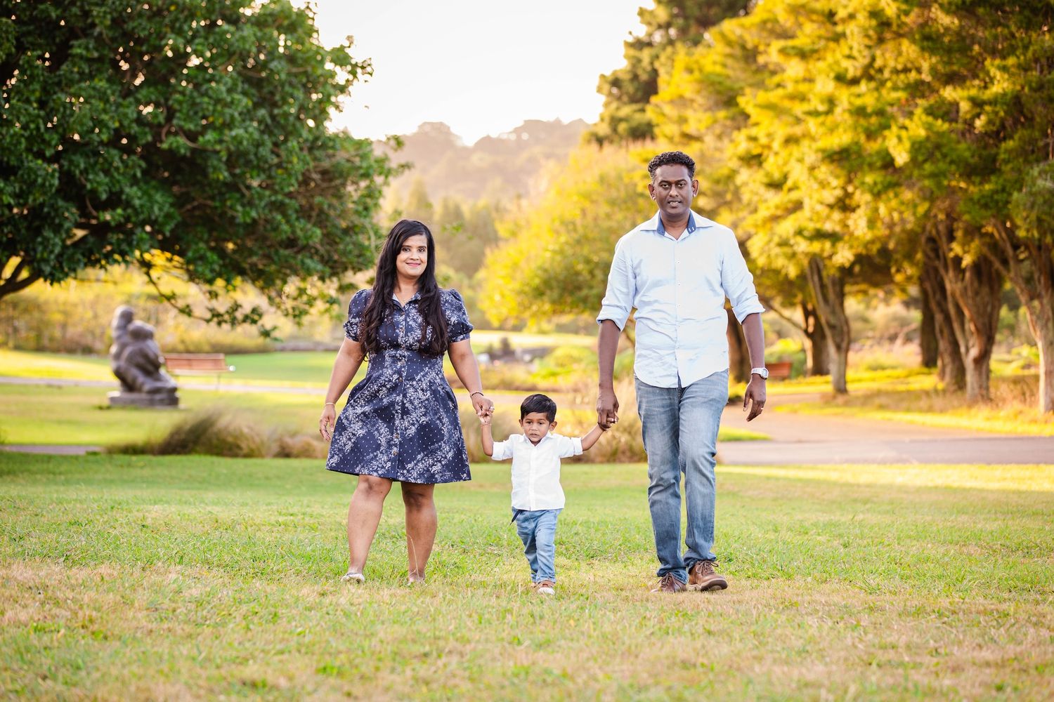 Family of three walking hand-in-hand through a lush green park during golden hour autumn season.