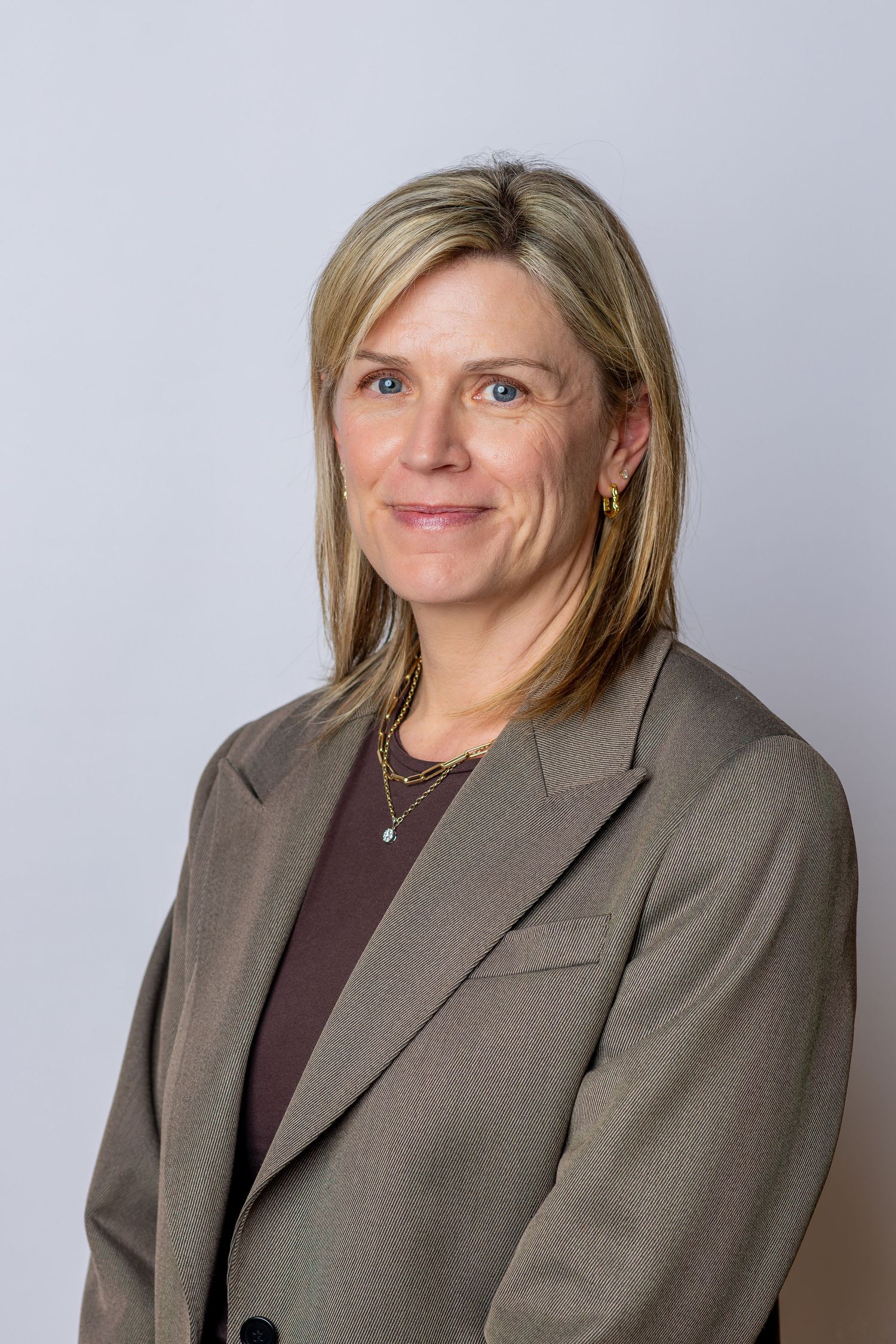 Professional headshot of a blonde woman in a gray blazer and gold jewelry smiling confidently against a light background.