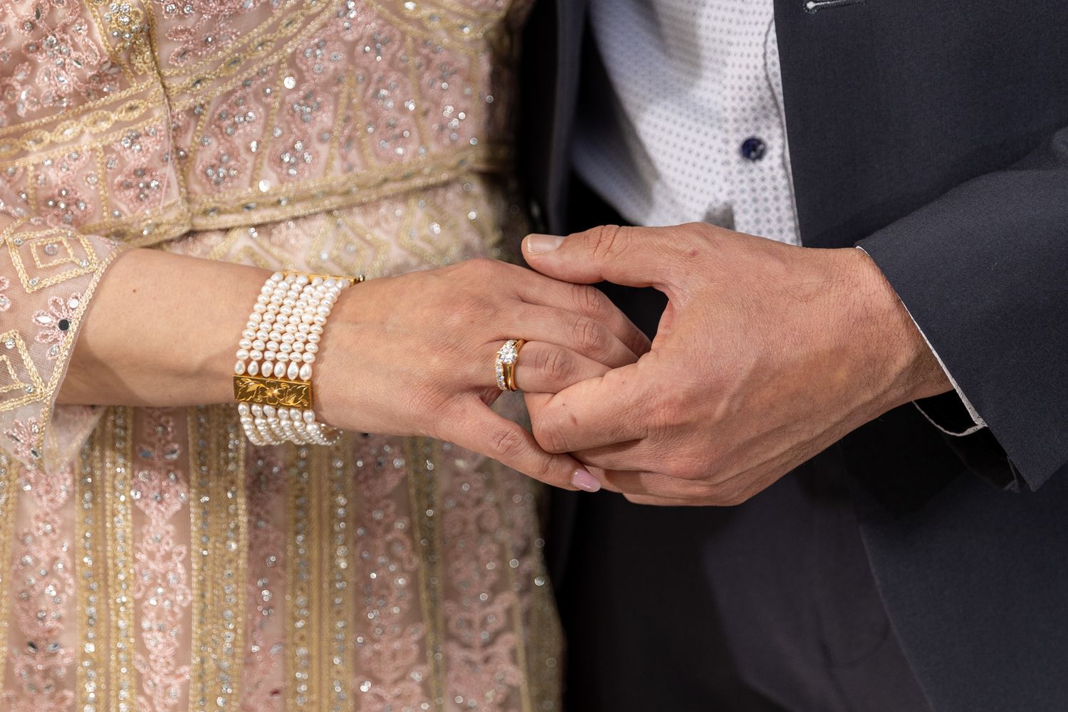 Close-up of couple holding hands, woman wearing pearl bracelet and ring, man in suit at wedding ceremony.