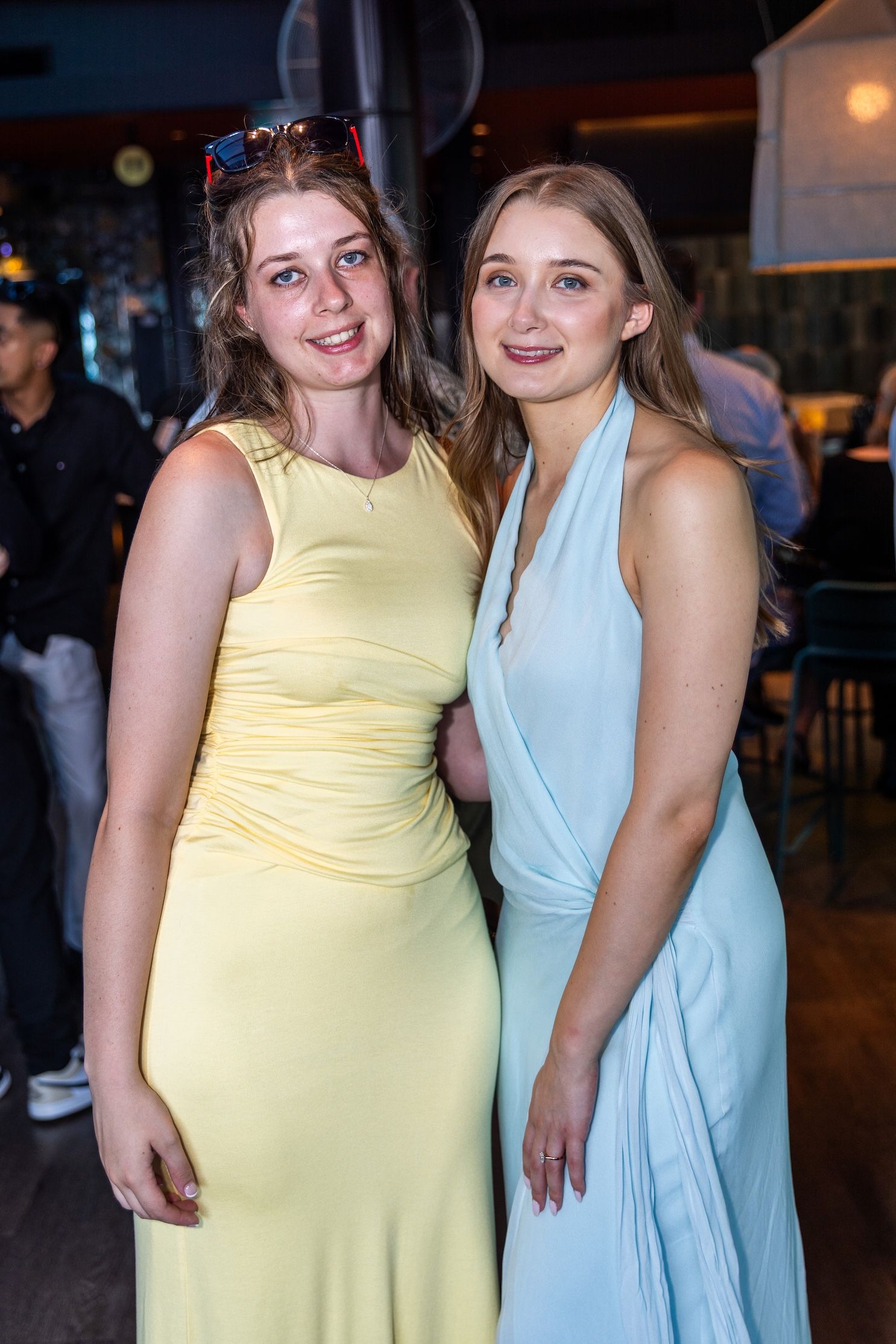 Two young women in elegant pastel formal gowns posing together at an indoor event venue.