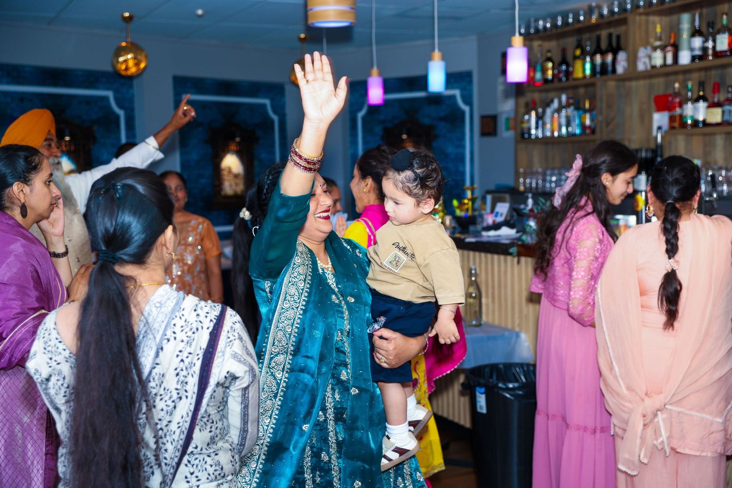 A woman in a teal sari holds a baby while dancing at a vibrant South Asian celebration inside a bar.