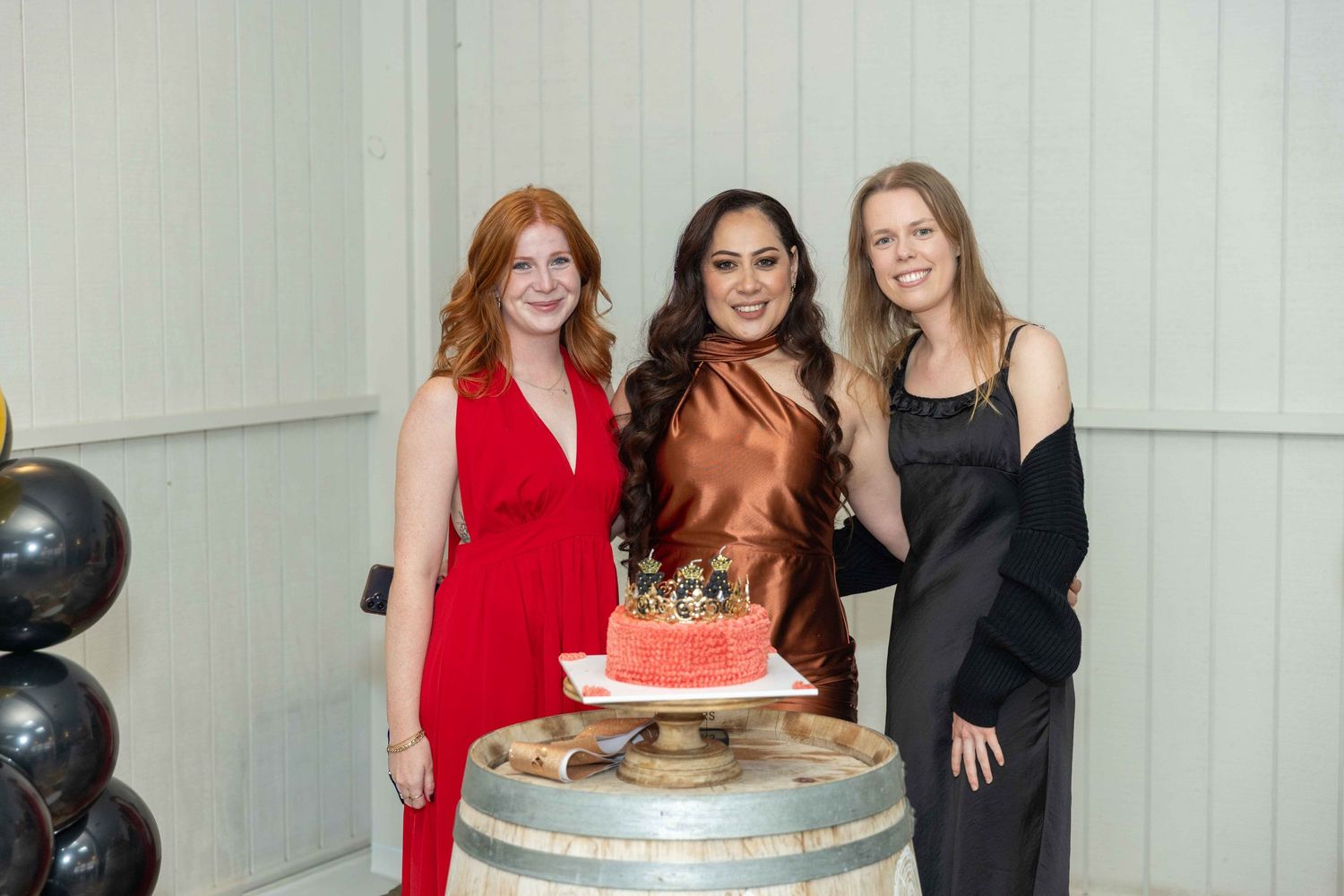 Three women posing with a pink birthday cake topped with a gold crown on a barrel at an elegant party.
