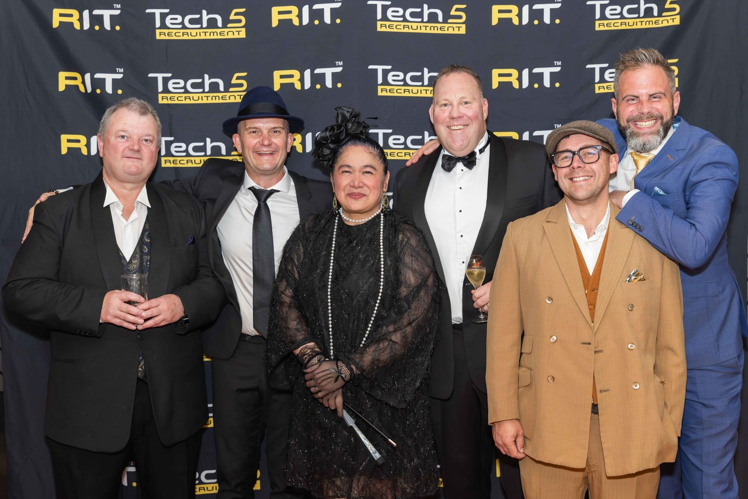 Six professionals pose together at an R.I.T. Tech5 awards event, dressed in formal and vintage-inspired attire.