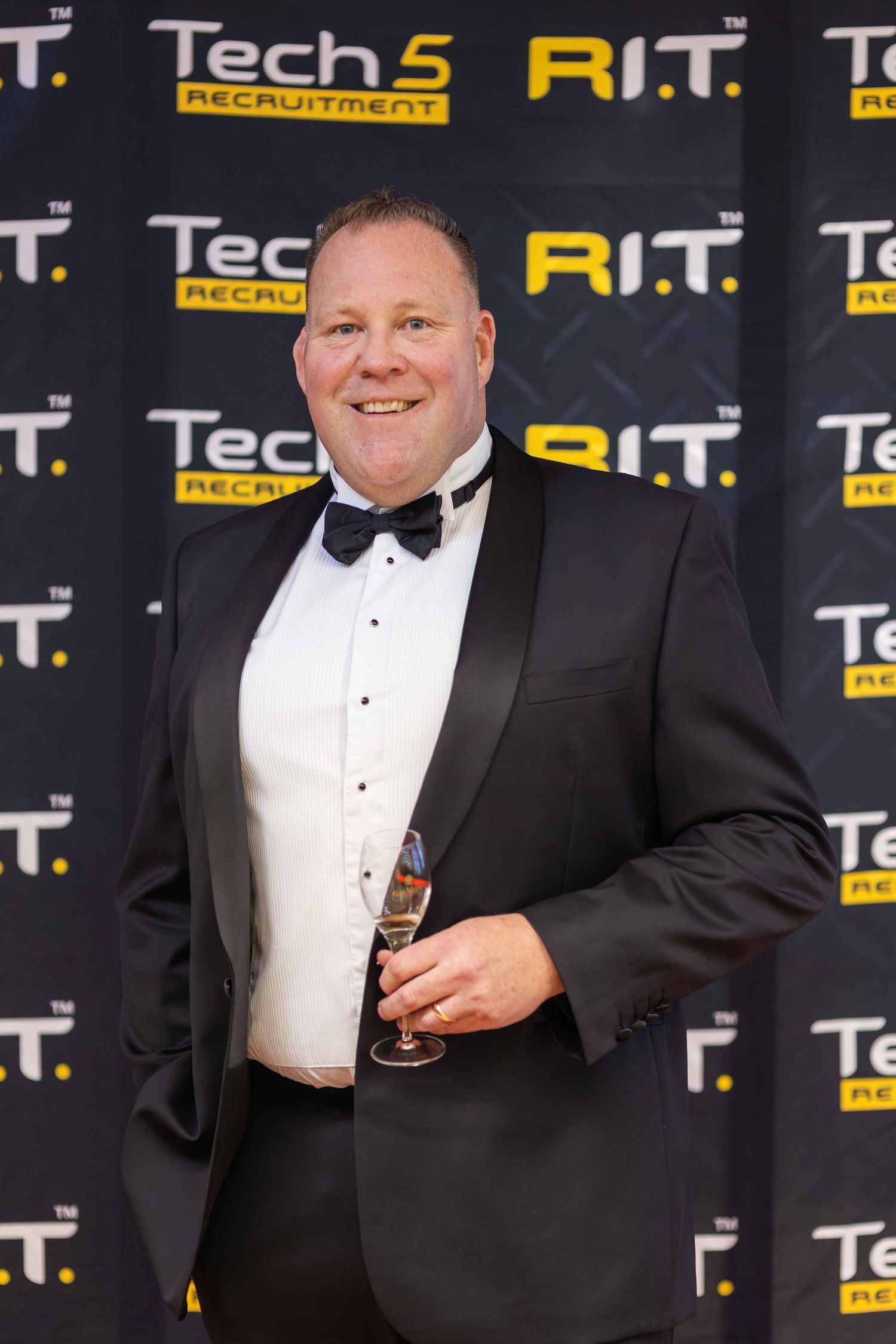 Man in tuxedo holding champagne glass poses at Tech 5 Recruitment and R.I.T. event backdrop.