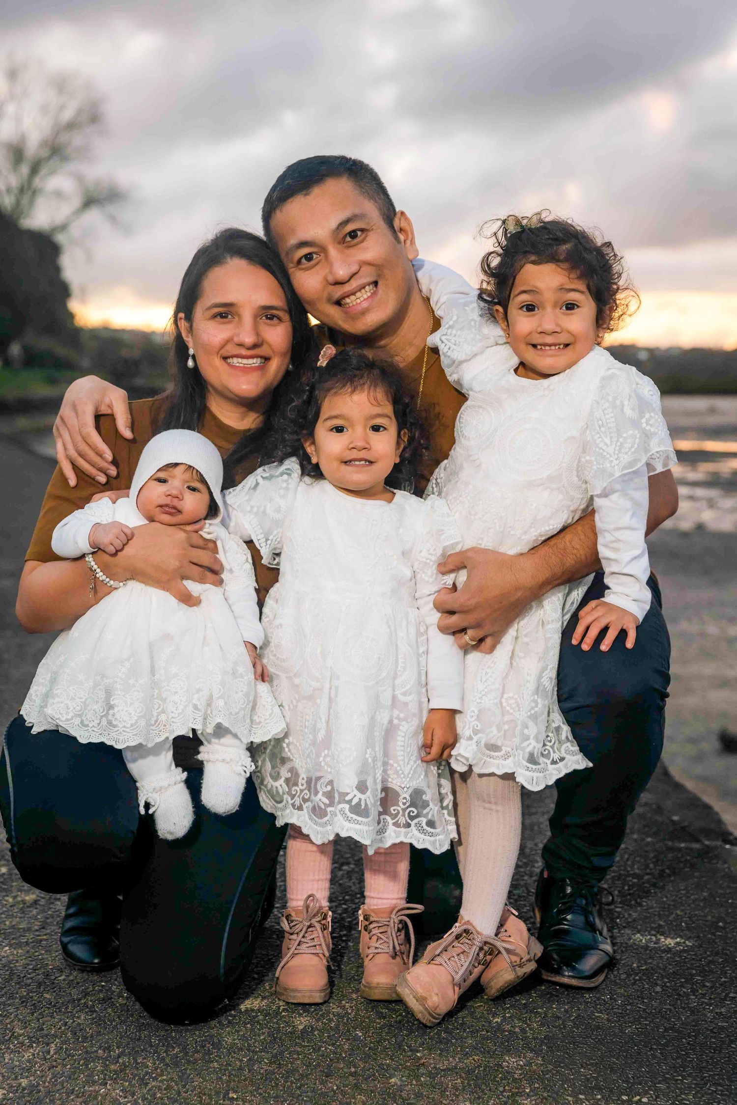 Happy family of five posing outdoors at sunset, parents with three young daughters in white dresses.