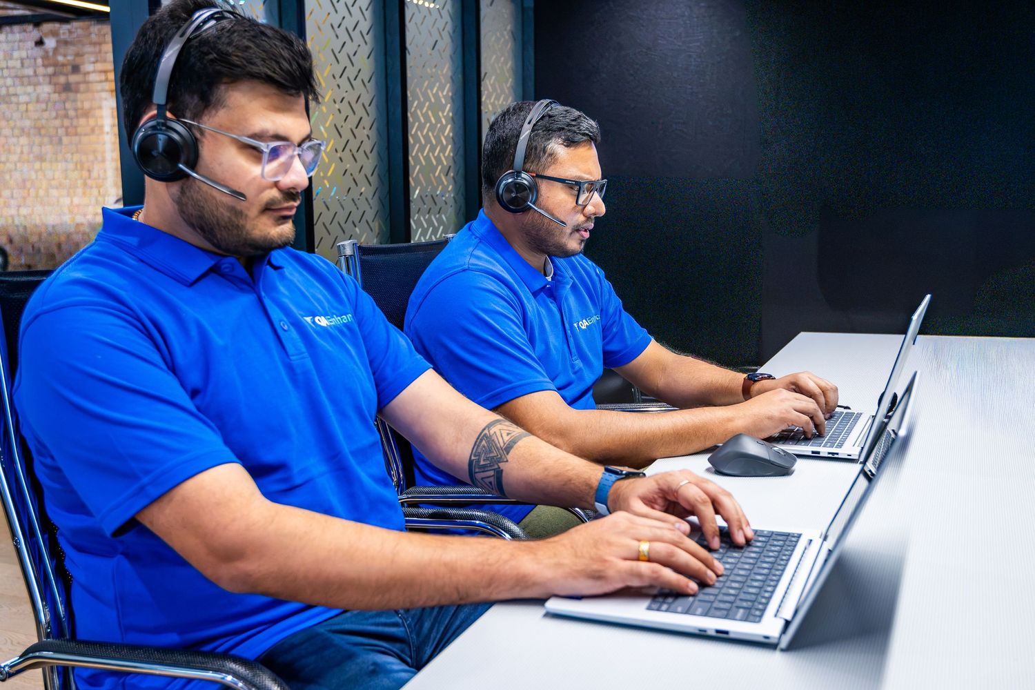 Two male call center agents wearing blue polo shirts and headsets working on laptops in a modern office.