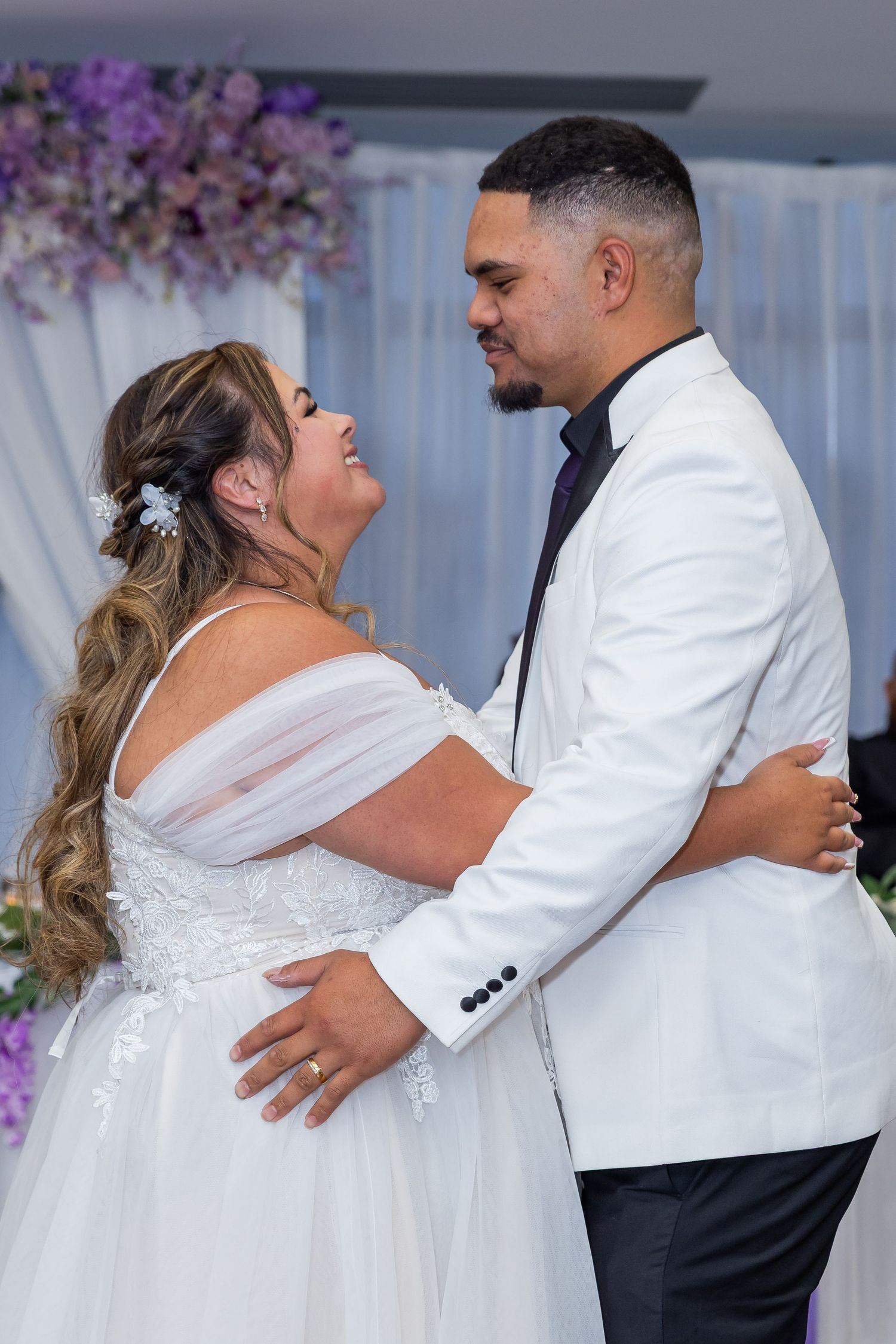 Bride and groom share first dance at wedding reception, bride in white gown, groom in white tuxedo jacket.