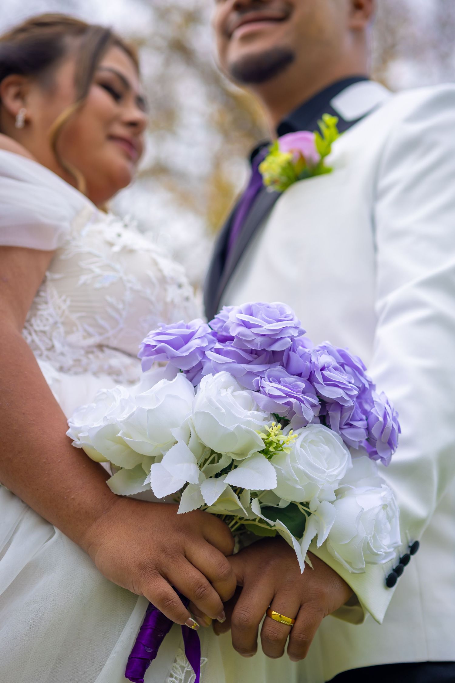 Bride and groom holding a bouquet of white and purple roses, with the groom wearing a gold wedding band.