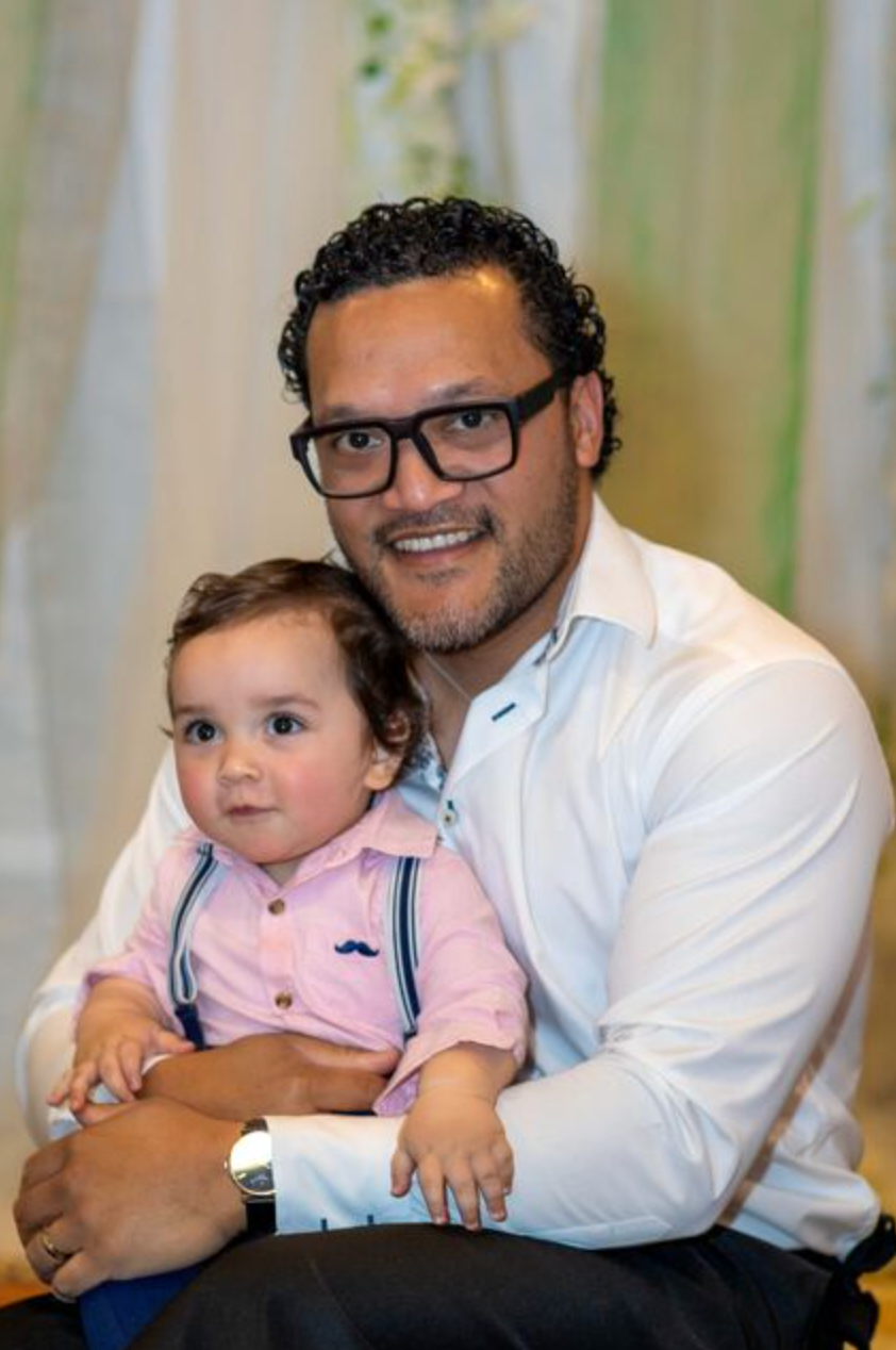 A smiling man with glasses in a white shirt holds a baby boy in a pink shirt with suspenders at a formal event.