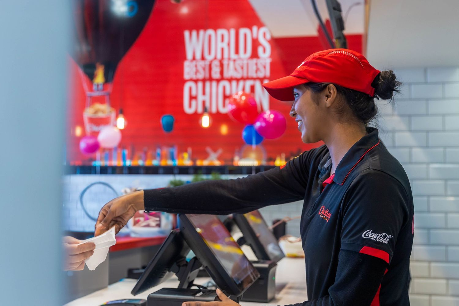 A smiling fast food employee in red cap and uniform works at a register under a World's Best Chicken sign.