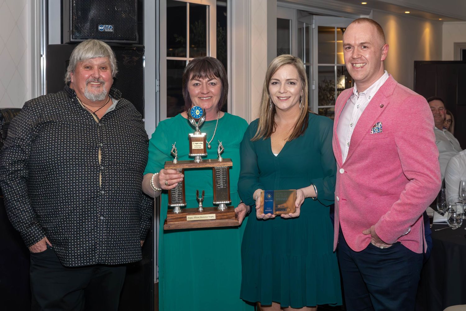 Four people posing with trophy and award at formal banquet event, wearing elegant attire in a restaurant setting.