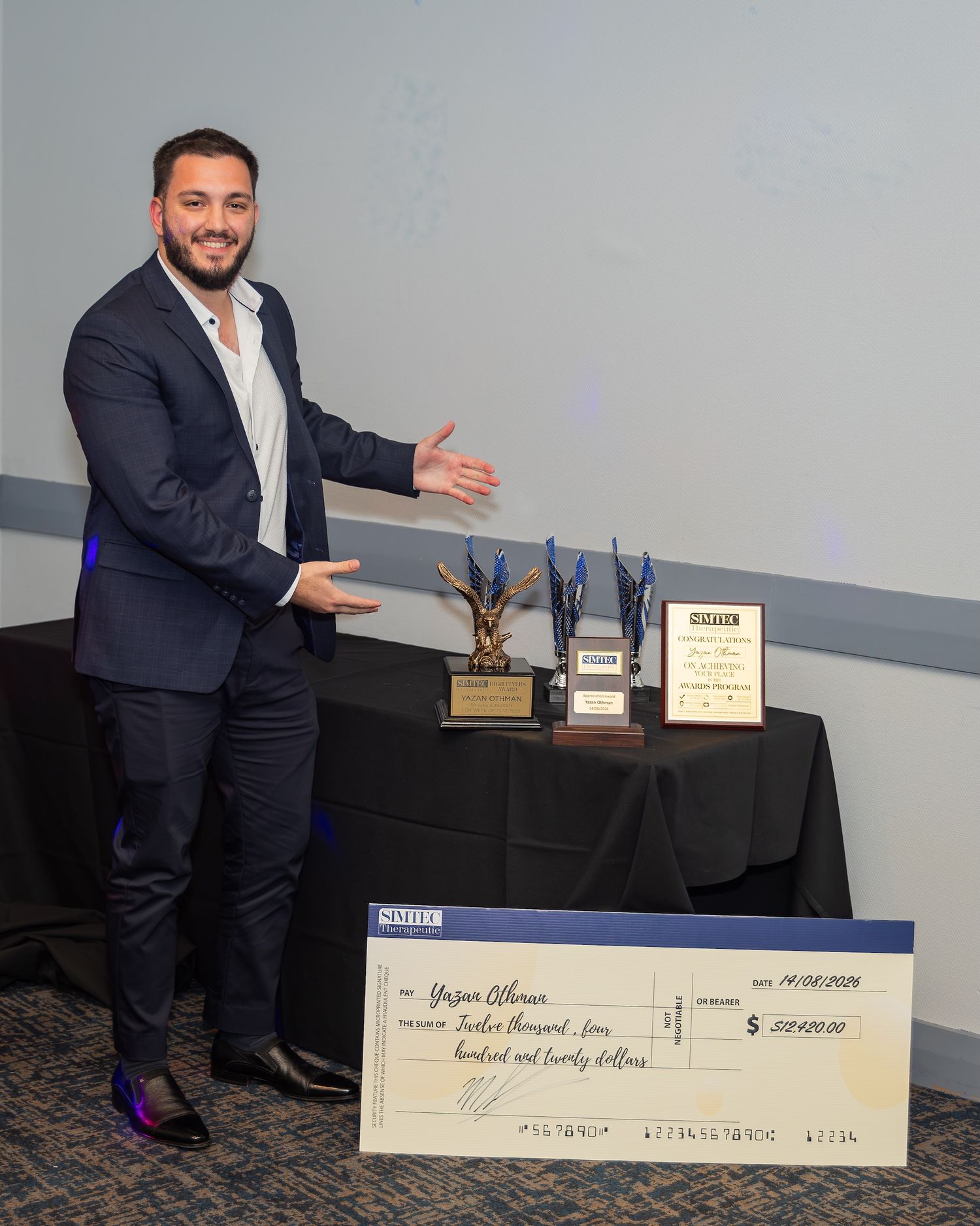 Man in suit smiling beside oversized check for $12,475 and trophy awards at a recognition ceremony.