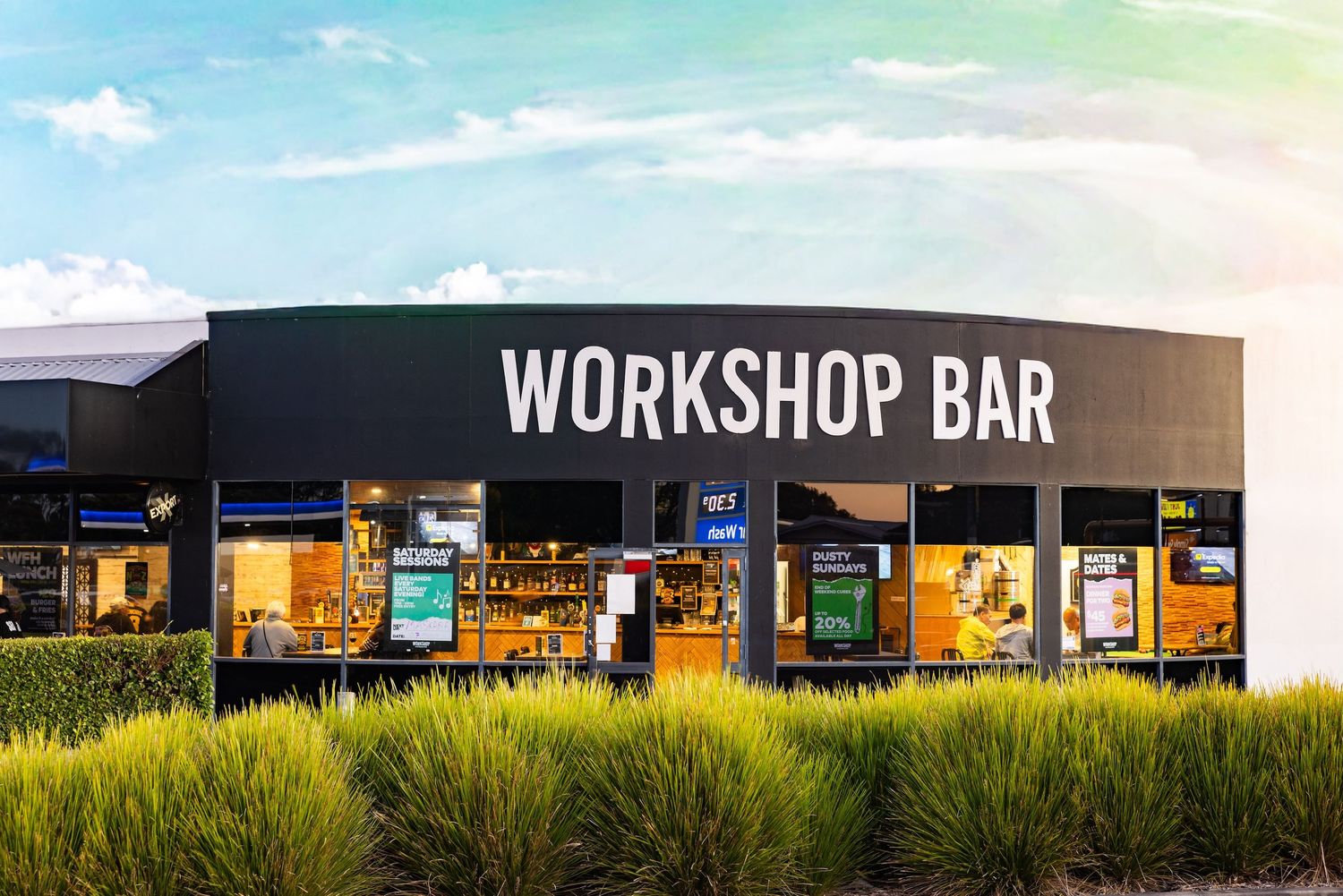 Modern black workshop bar building with illuminated signage and glass windows set against a natural grass landscape at dusk.