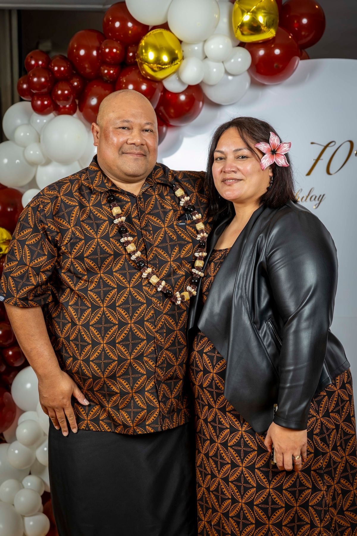 Couple poses in front of balloon arch backdrop wearing coordinated brown patterned and black formal attire.
