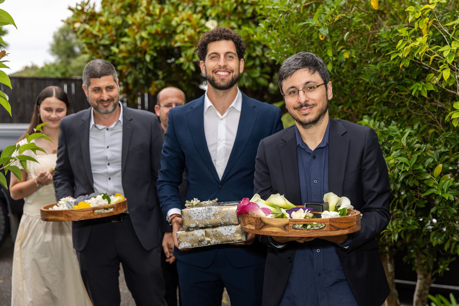 Men in suits carry decorative trays with flowers and food outdoors at a formal garden event.