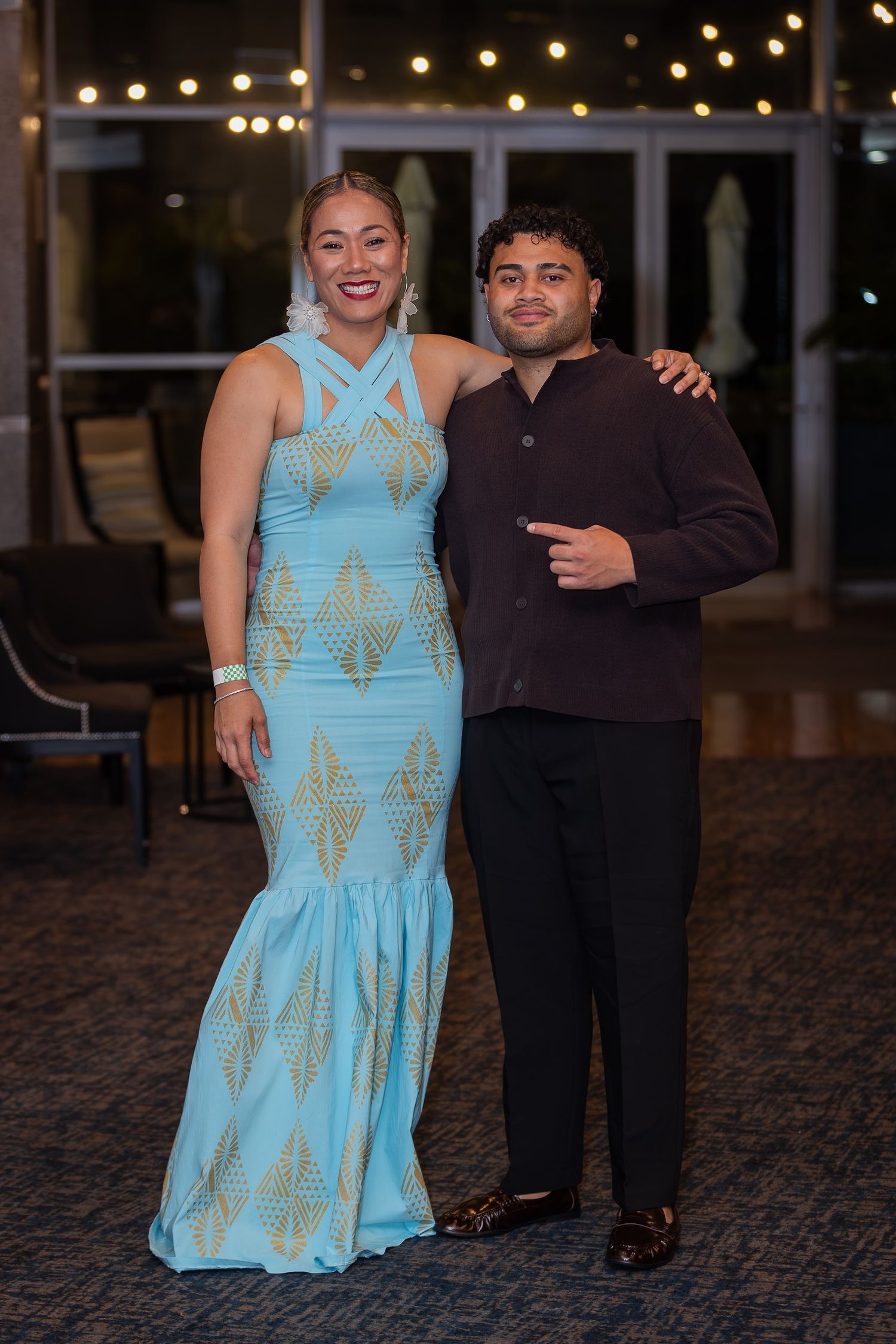 A couple posing at prom, woman in light blue gold gown, man in dark suit, string lights in background.