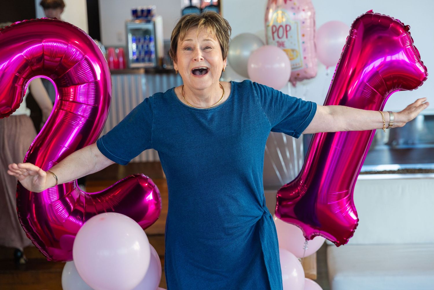 Woman celebrating 21st birthday with pink foil balloons, laughing and posing with arms outstretched at a party.