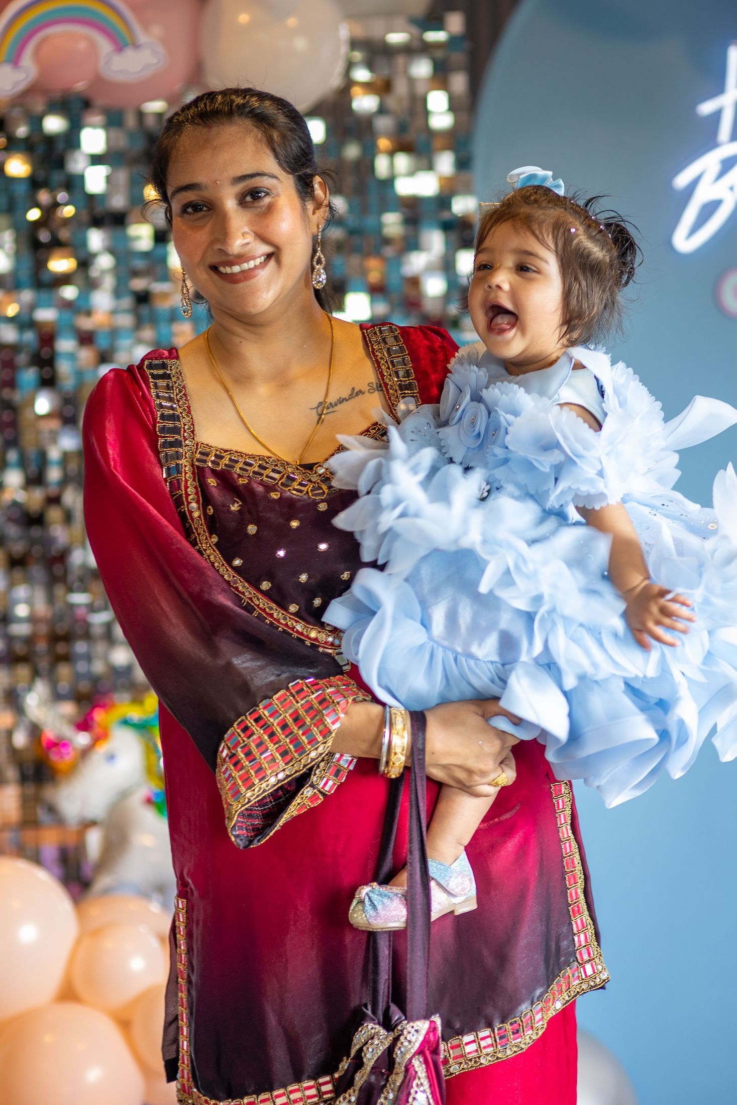 A smiling woman in a red traditional Indian outfit holds a baby girl in a light blue ruffled dress at a birthday party.