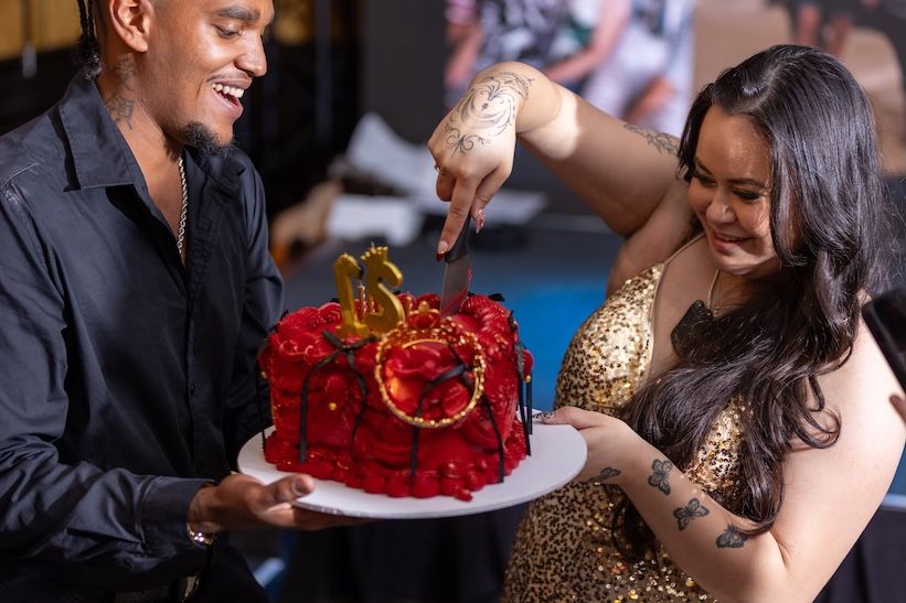 A man and woman cutting a red rose birthday cake decorated with gold numbers at a celebration.