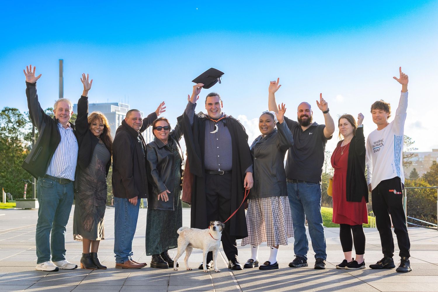 Graduate in cap and gown celebrates with family and friends outdoors, all cheering with arms raised.