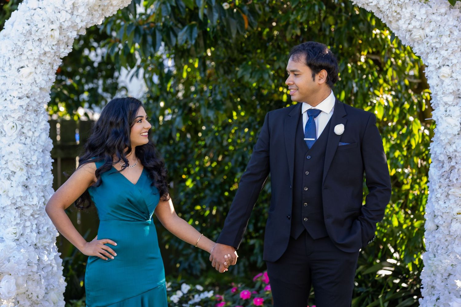 Couple holding hands under a white floral arch, woman in teal dress, man in dark suit, outdoor garden setting.