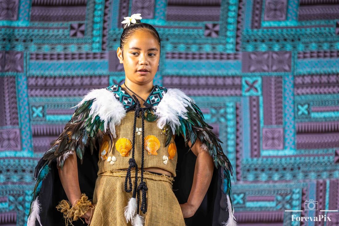 Young woman in traditional Polynesian costume with feathered cape performing on stage with tapa cloth backdrop.