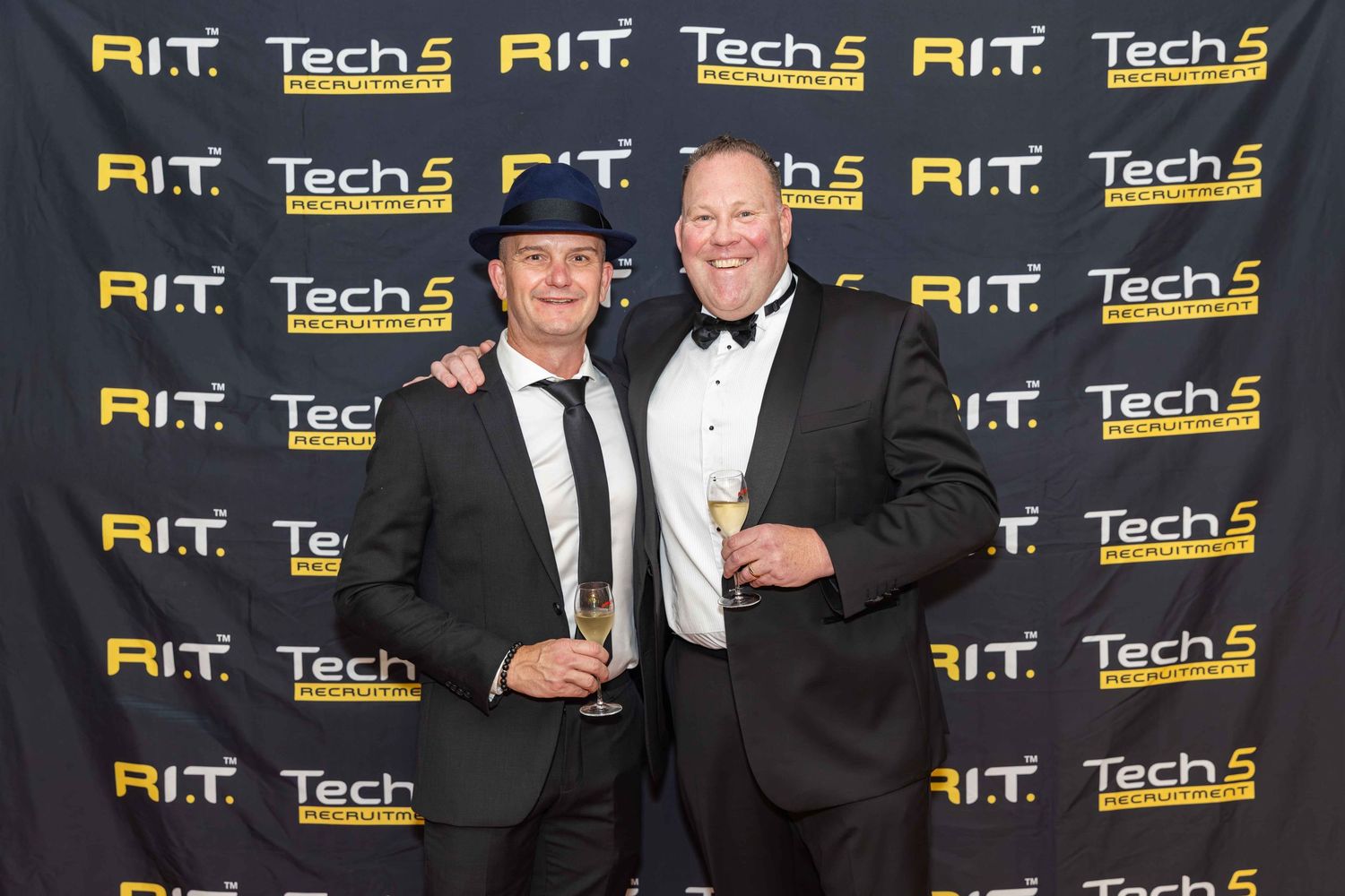 Two men posing at RIT Tech 5 Awards event, one wearing a hat and suit, the other in a tuxedo, holding champagne glasses.