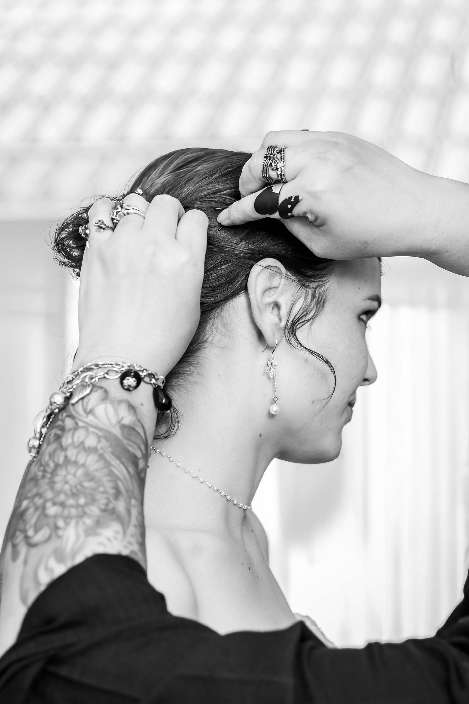 Black and white photo of tattooed hands styling a bride's updo, adorned with rings and bracelet.