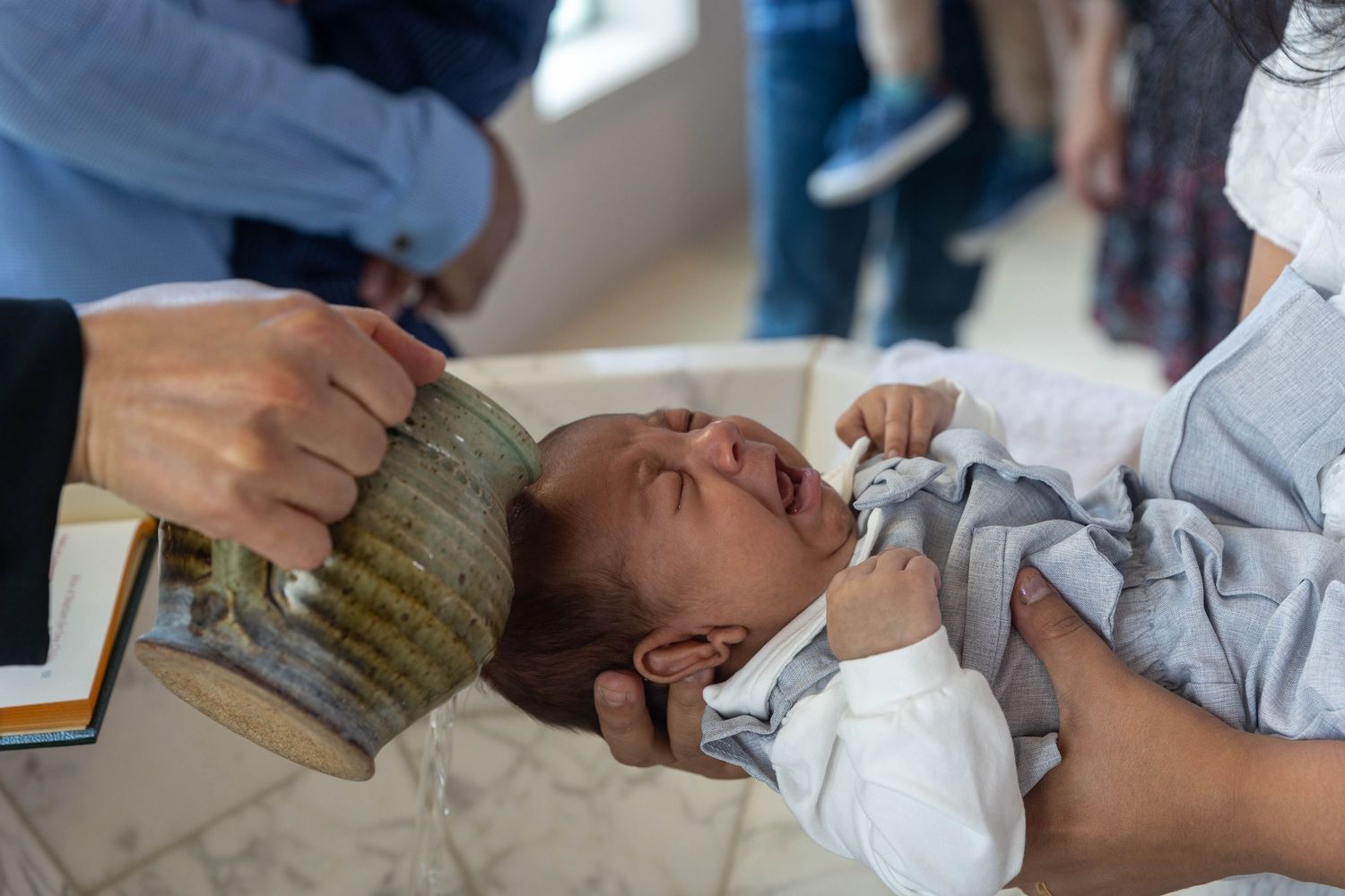 A crying newborn baby being baptized with water poured from a ceramic vessel during a Christian baptism ceremony.