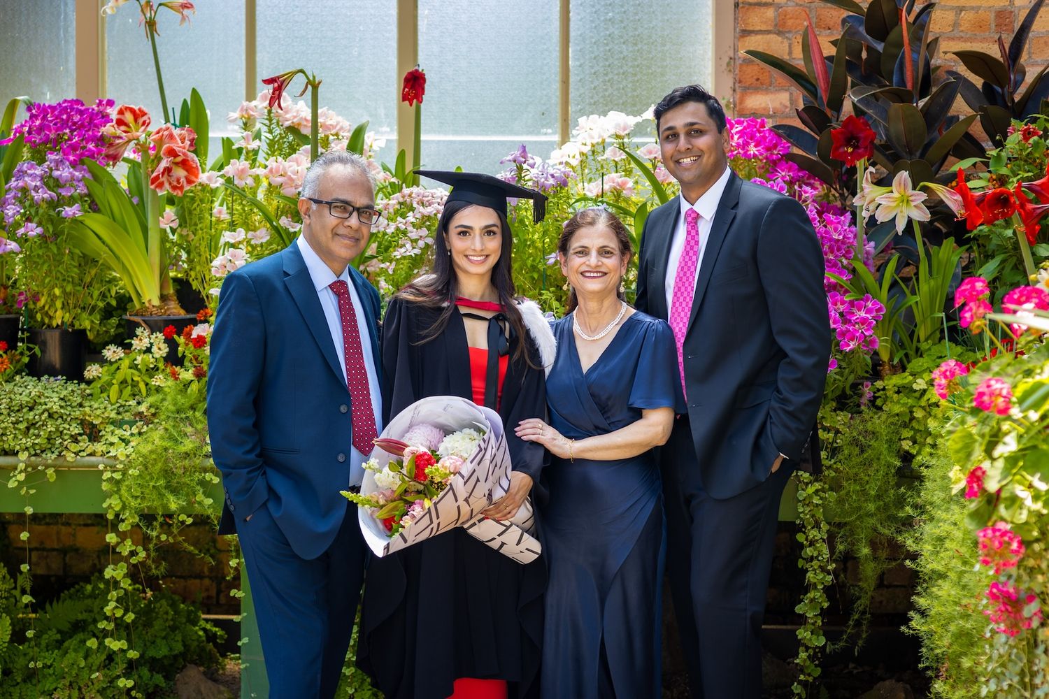 Graduate in cap and gown poses with family among colorful flowers, holding a bouquet to celebrate her graduation.