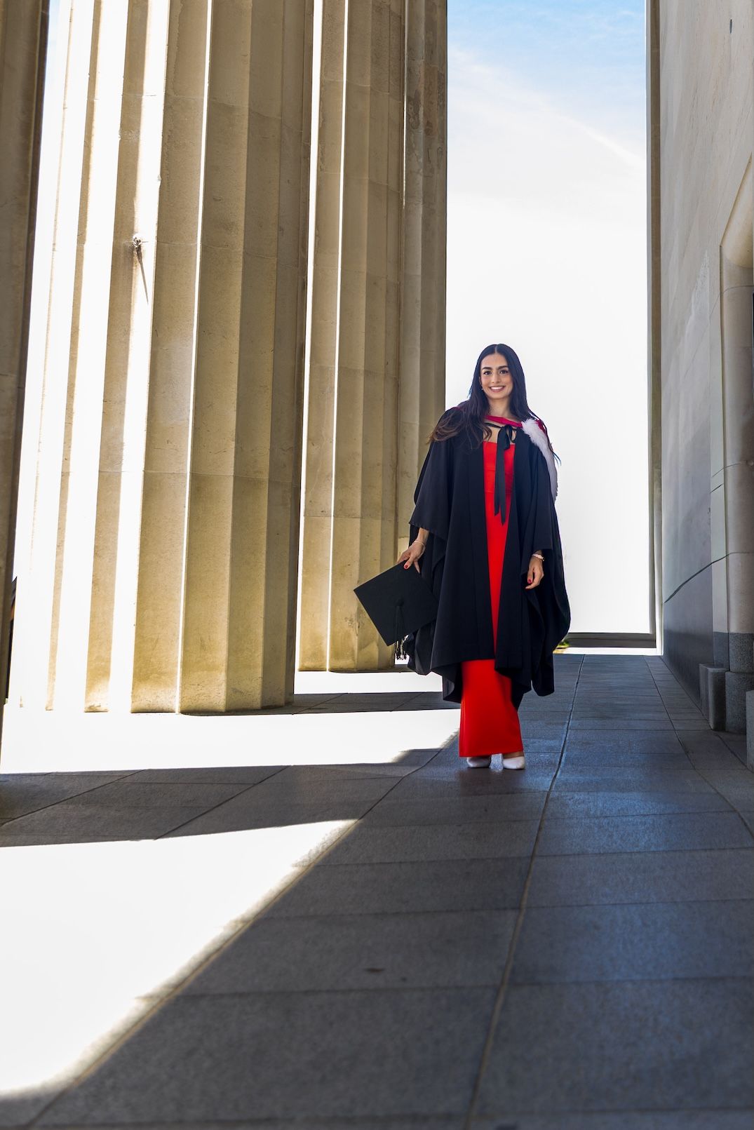 A smiling graduate in black gown and red dress walks between tall stone columns on graduation day.