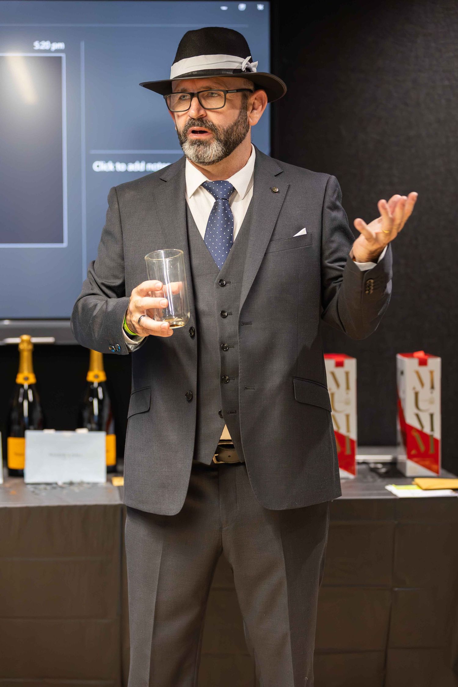 Elegant man in gray suit and black fedora holds glass while gesturing at upscale beverage presentation event.