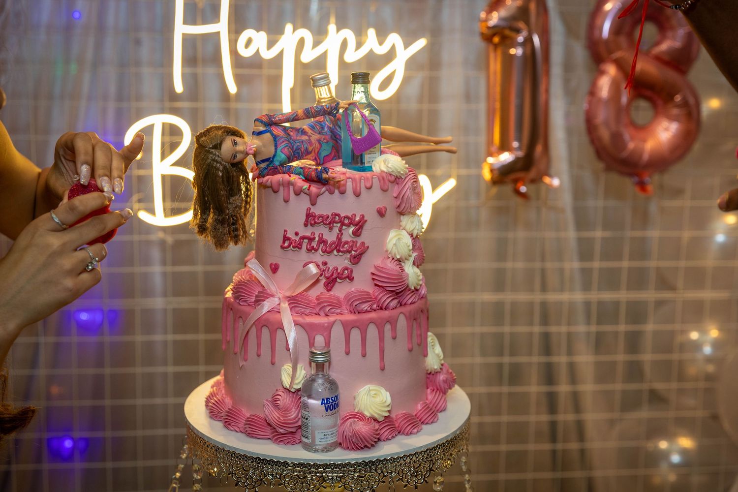 Pink two-tier birthday cake with happy birthday Siya decoration, neon sign backdrop, and balloon decorations at a birthday party.