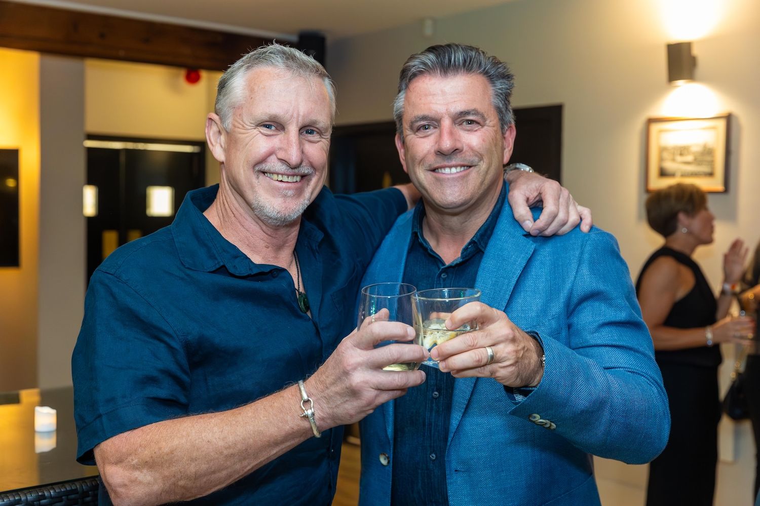 Two smiling men in blue attire toasting drinks at an indoor social gathering event.