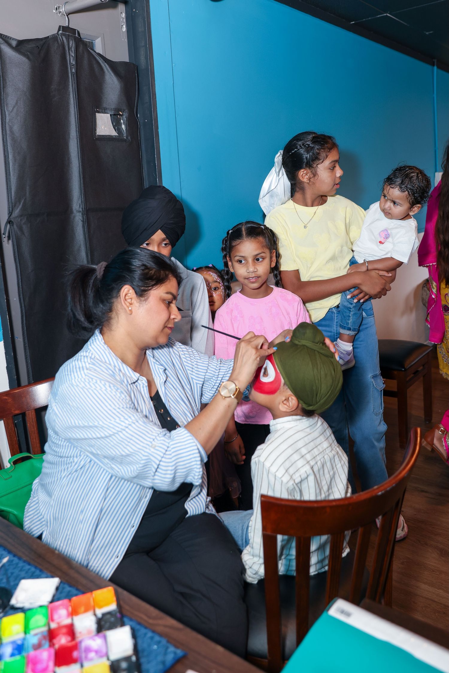 A woman paints a child's face at a party while other children and adults watch in a colorful indoor setting.